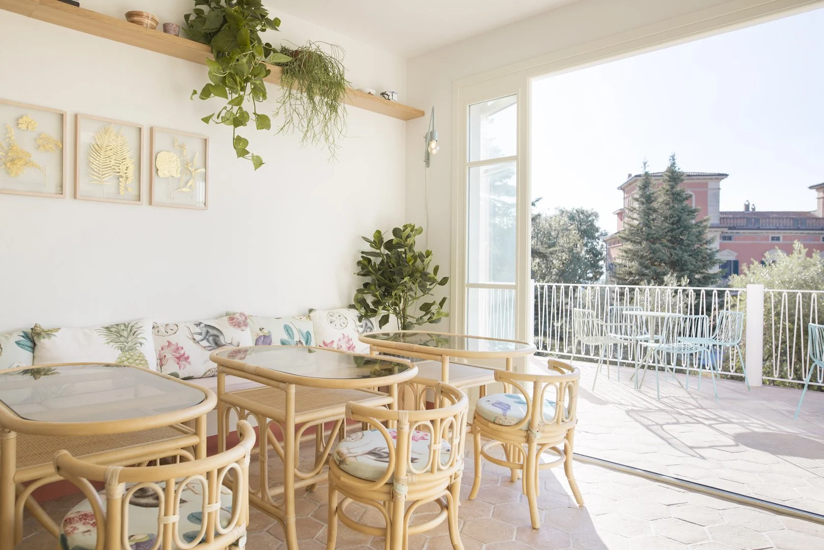 Bright indoor dining area with rattan furniture and floral cushions, open to a balcony with outdoor tables and chairs, greenery outside, and decorative wall art.