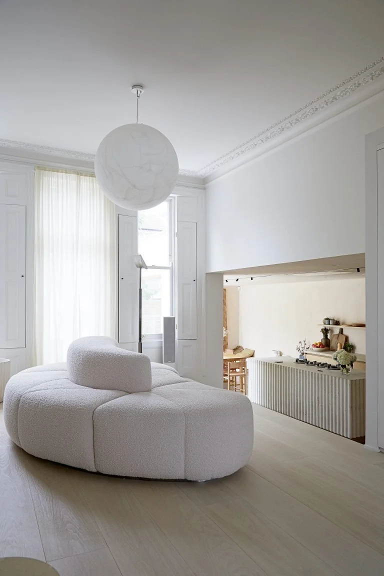 Bright, minimalist living room with white rounded sofa, large windows with sheer curtains, a hanging spherical white light fixture, and a partial view of a kitchen or dining area with a striped counter.