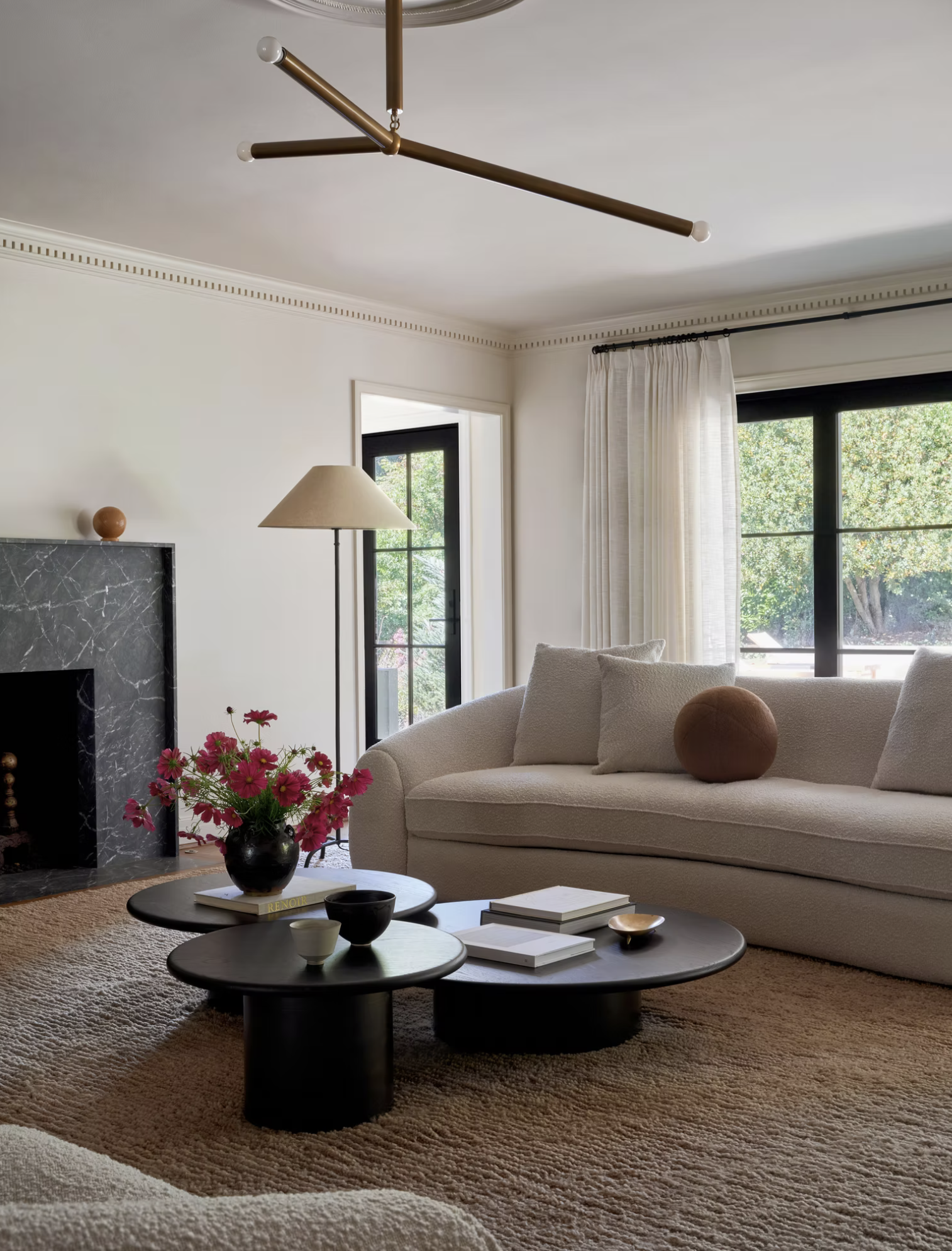 Living room with beige sofa, black coffee tables, vase of pink flowers, floor lamp, fireplace with black marble surround, large window with white curtains, beige carpet, and modern ceiling light fixture.