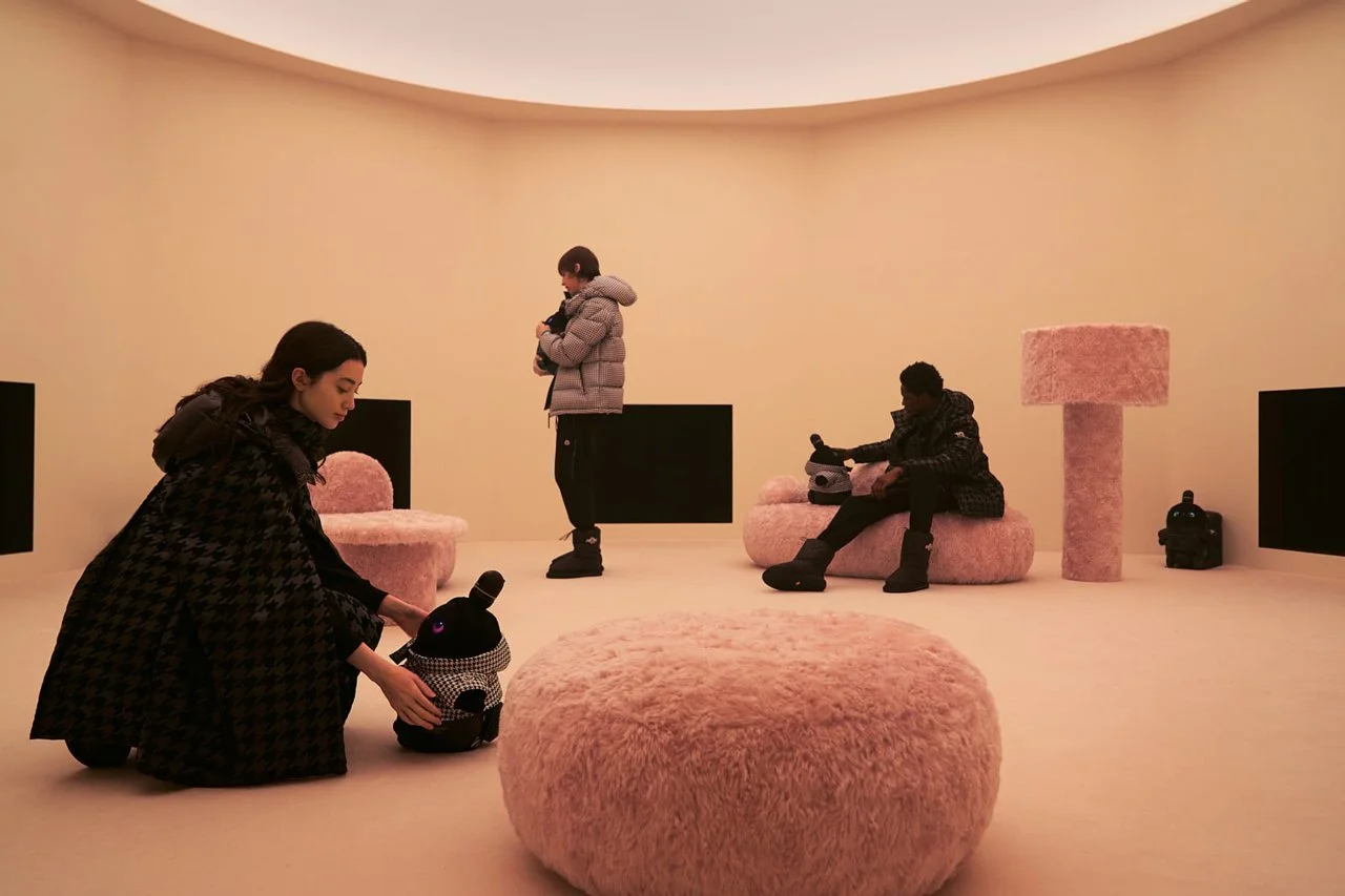 Three women in a minimalistic, pink decor room with plush furniture. One woman is kneeling, adjusting a plush bunny-shaped speaker. Another woman is standing, looking at her phone, while the third woman is sitting on a pink covered sofa with a helmet
