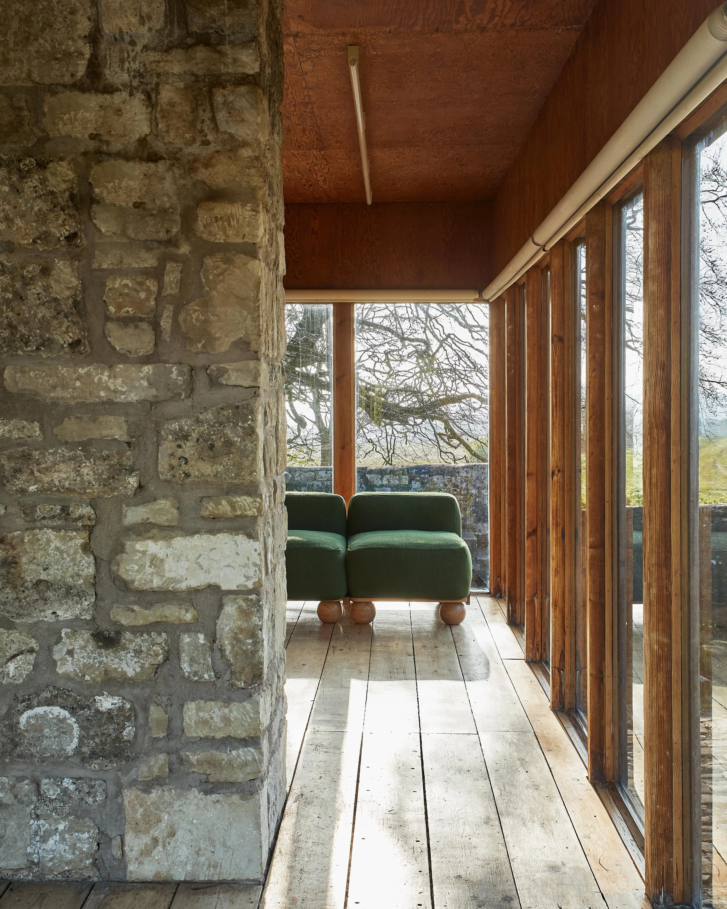 Interior view of a rustic room with a stone column, wooden ceiling, large windows, and a green sofa with wooden legs, overlooking a scenic outdoor view with trees.
