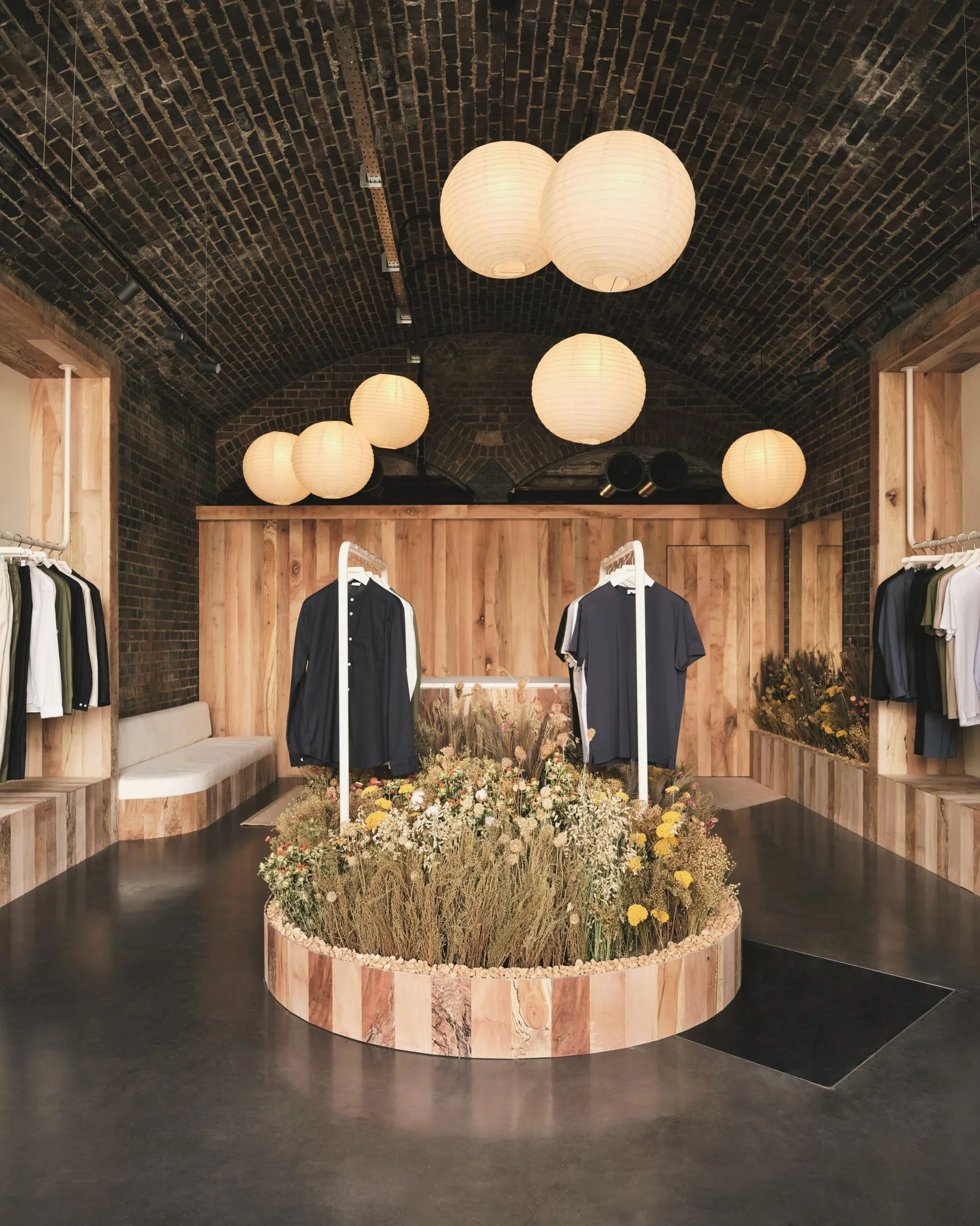 A clothing store with a central circular flower bed filled with various flowers and plants. Wooden walls and black flooring, black brick arched ceiling, hanging round paper lantern lights, racks of men's and women's clothing on both sides.