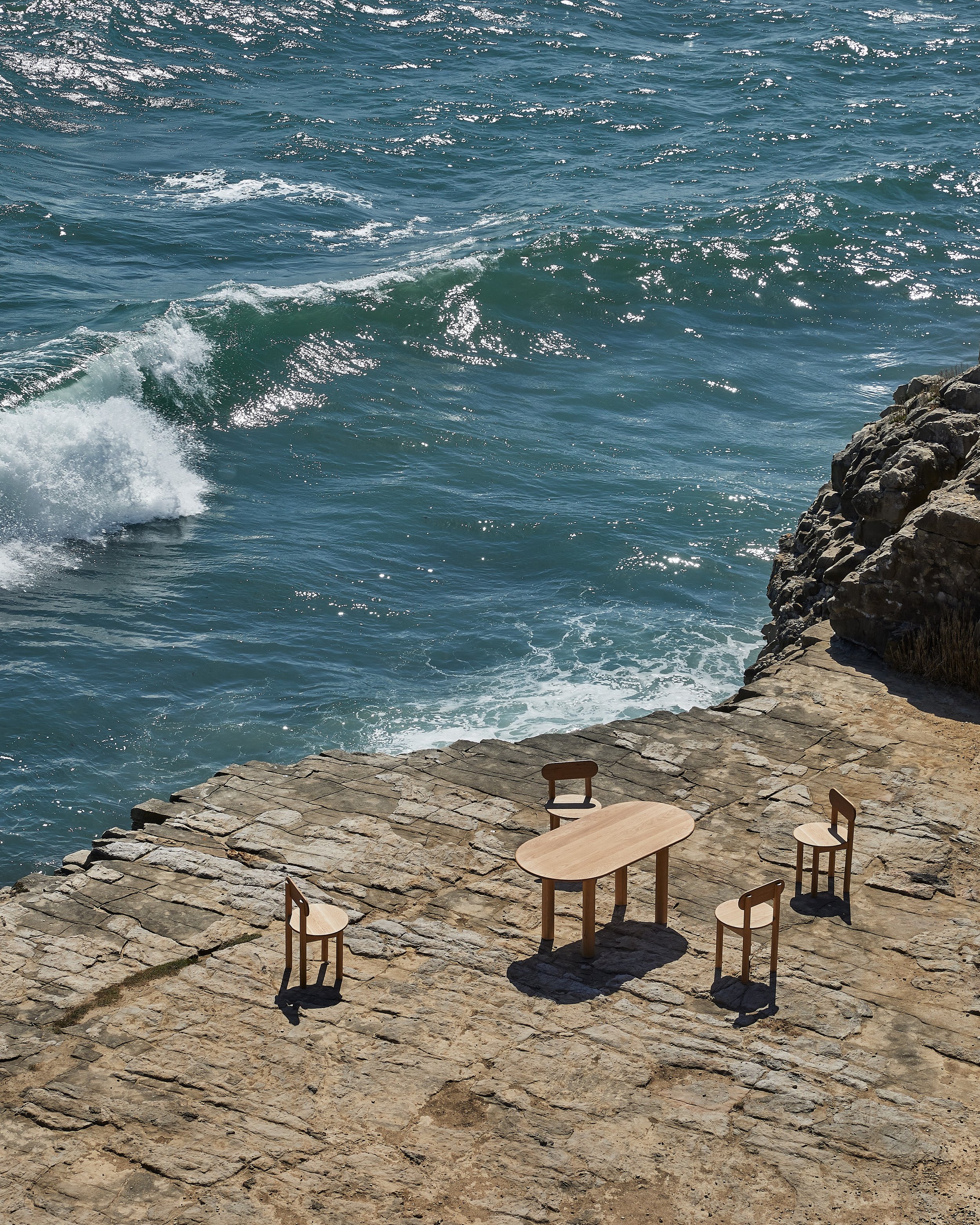A rocky seaside area with a small wooden table and four chairs, overlooking the ocean with waves crashing against the rocks.