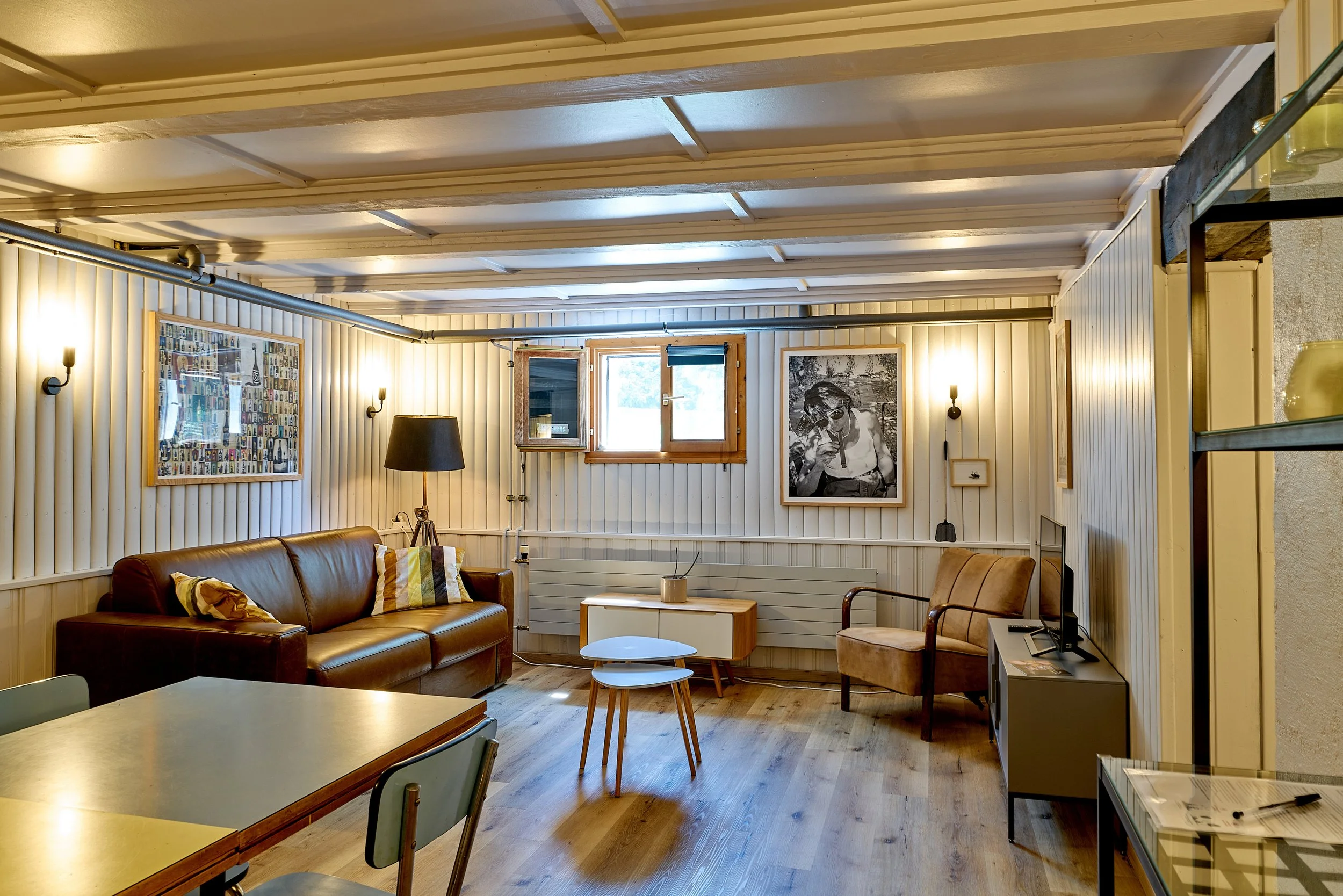 Salon cosy avec un canapé en cuir marron, wifi, une table basse, un fauteuil en tissu beige, une télévision, des étagères en verre, des tableaux au mur (portrait photo original de Jacques Dutronc) des lampes, des fenêtres ouvertes.