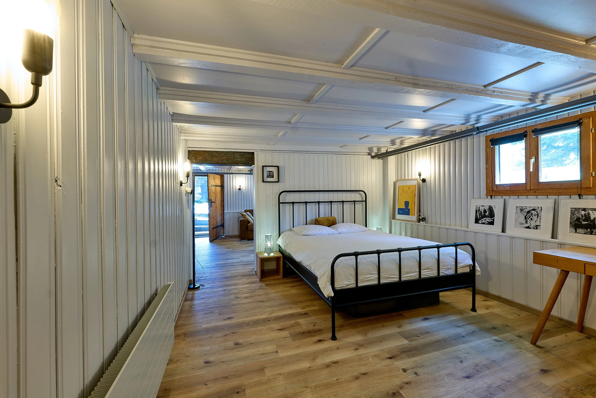 Chambre spacieuse avec lit en métal noir, murs en bois blanc, Une fenêtre donnant sur le jardin, parquet, décor moderne avec photos originales noir et blanc, lampes murales, porte en bois menant au salon séjour, ambiance chaleureuse, cocooning.