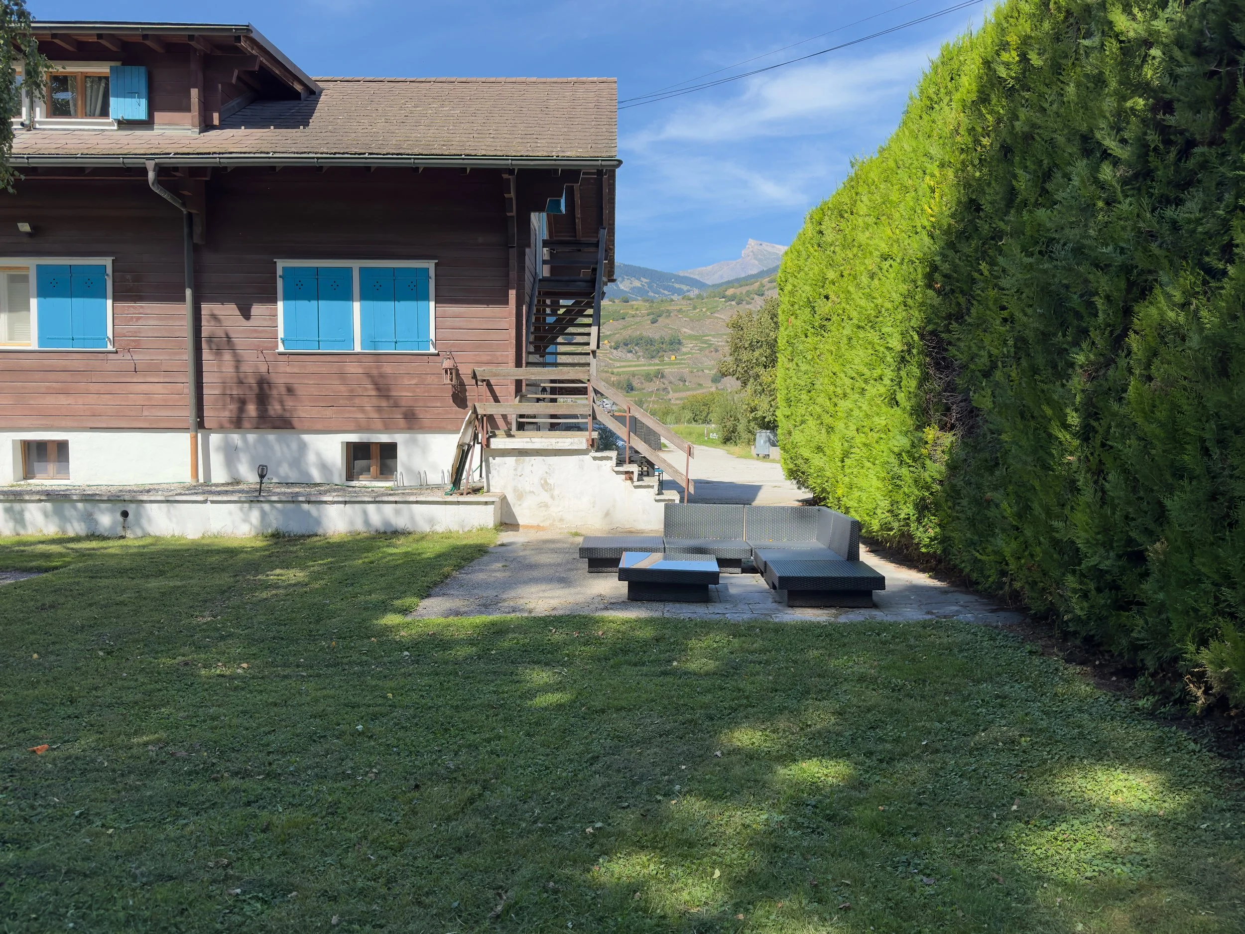 Maison en bois avec fenêtres à volets bleus, petits escaliers, espace de détente avec canapé lounge de jardin et table, grande haie verte côté droit, pelouse verte, paysage montagneux alpin en arrière-plan.
