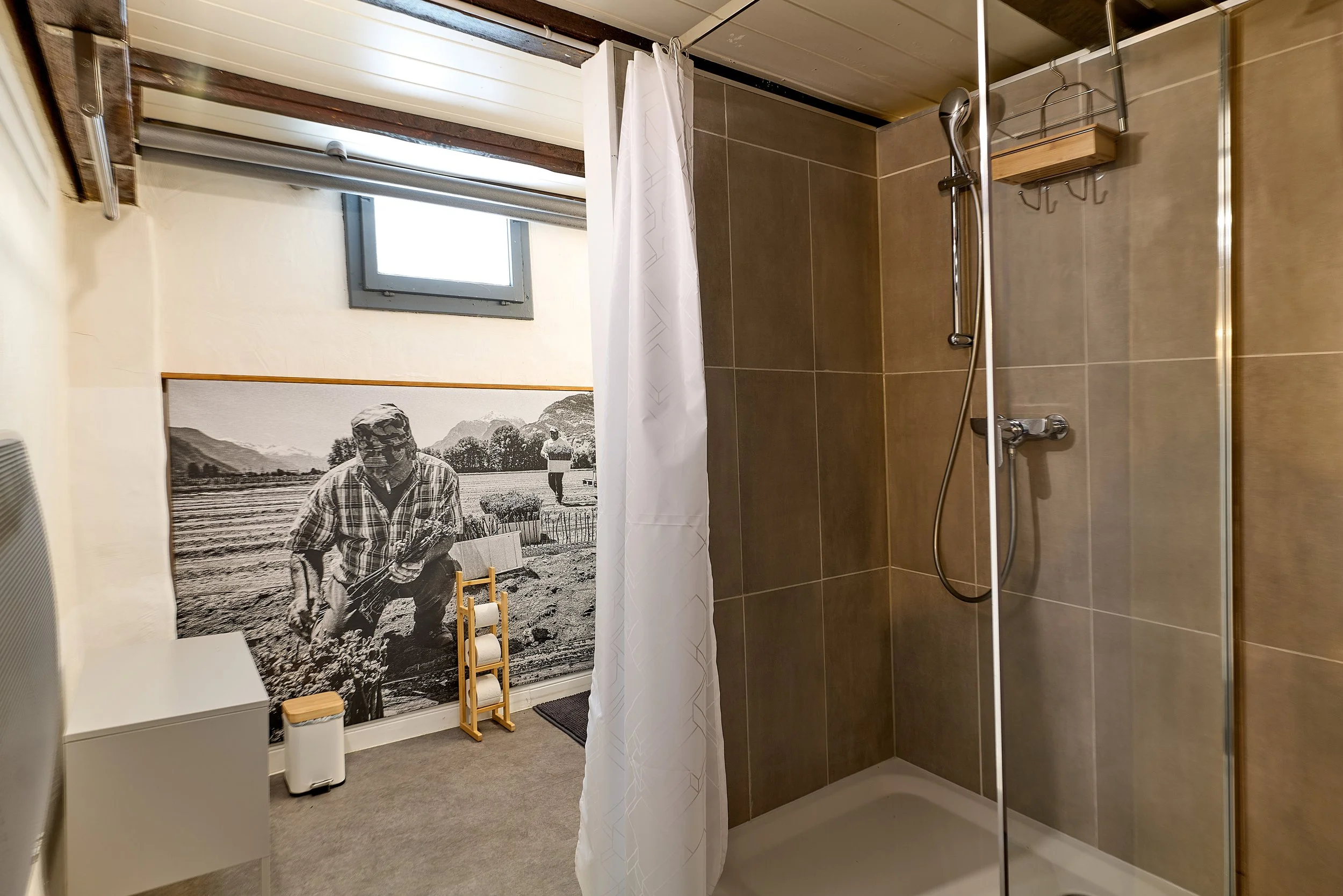 Salle de bain avec douche séparée, mur en carrelage gris, fenêtre donnant sur le jardin, mur décoré avec une grande photo noir et blanc d'un homme travaillant dans un champs.