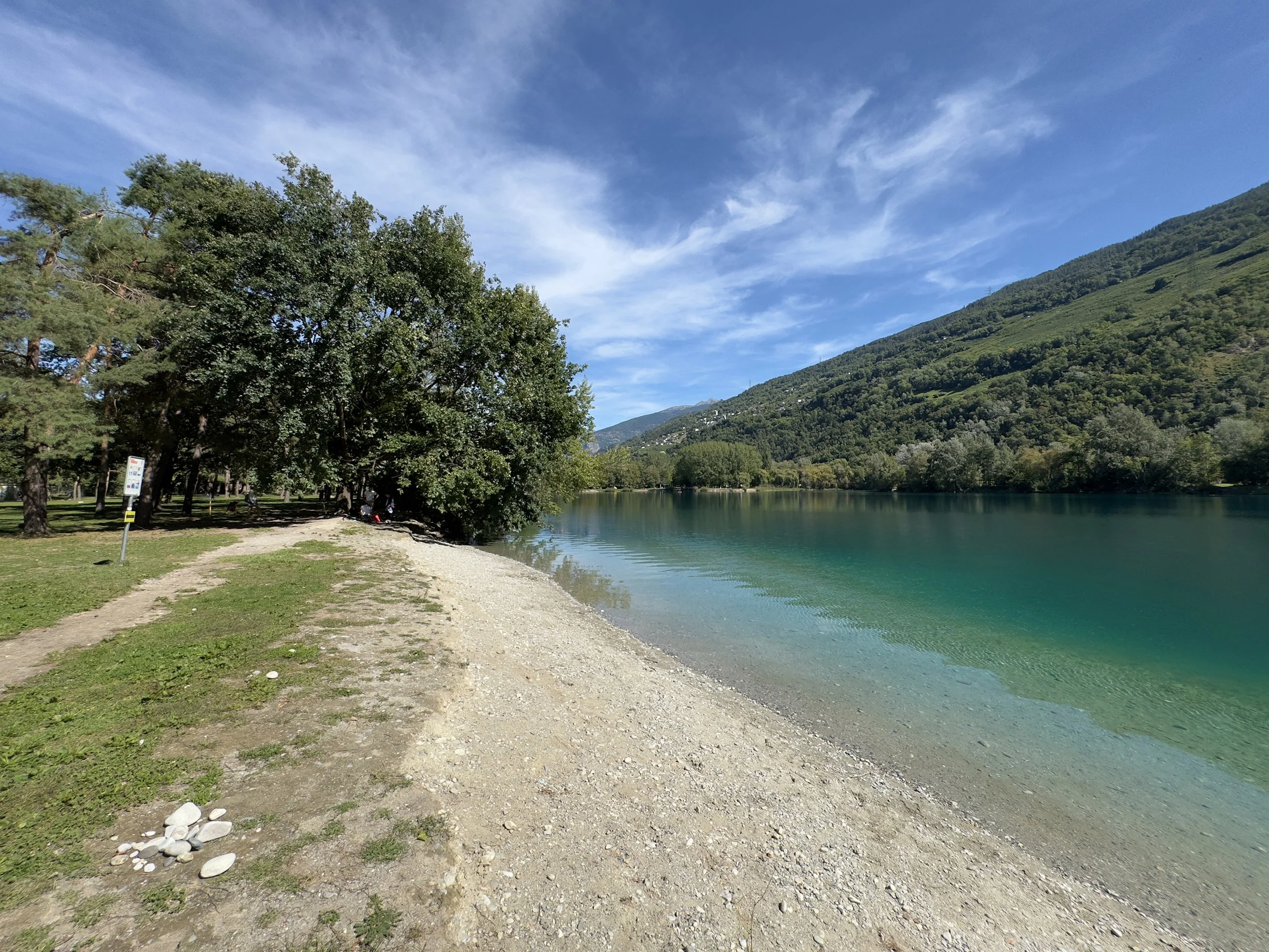 Au Domaine des Îles de Sion, en Valais (Suisse). Une plage de galets au bord d'un lac turquoise entouré de végétation et de montagnes, avec un ciel bleu et quelques nuages. Plaine, Valais romand.