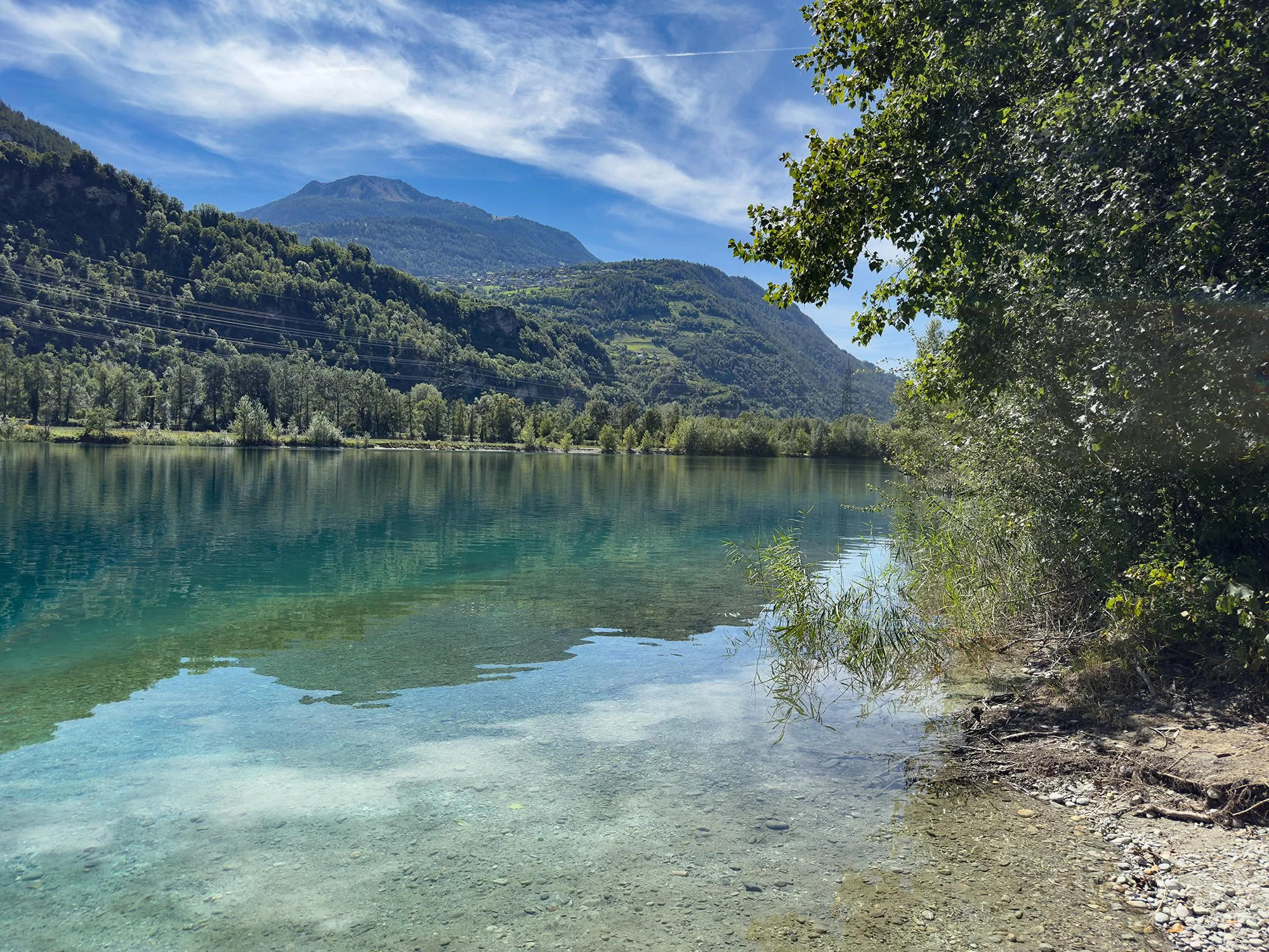 Lac du Domaine des Îles à Sion, en Valais. Parc nature et loisirs. Lac clair entouré d'arbres avec des montagnes en arrière-plan sous un ciel bleu avec des nuages wispy. Paysage alpin, Nendaz.