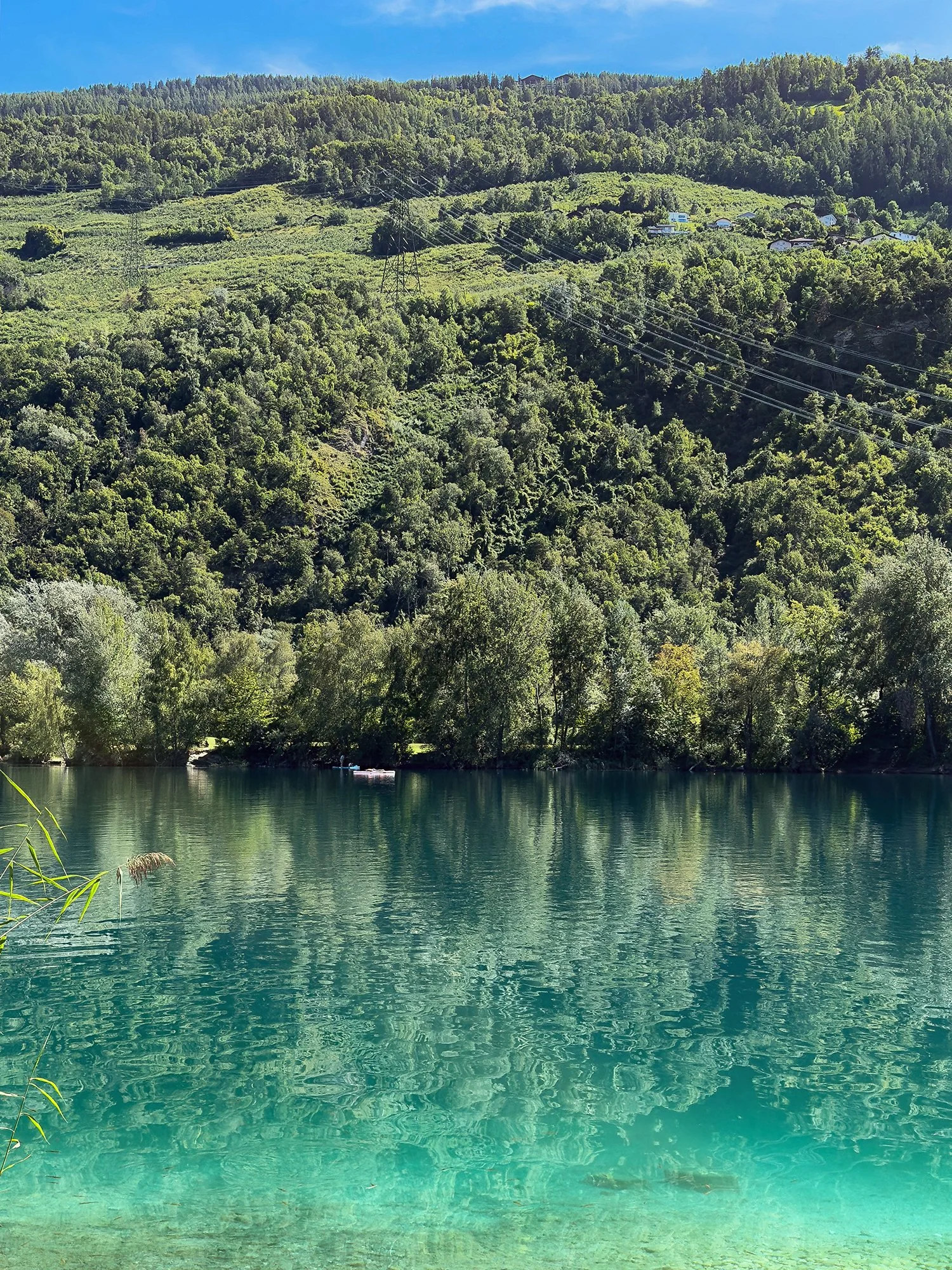 Au Domaine des Îles de Sion, en Valais (Suisse). lac turquoise entouré de végétation et de montagnes, avec un ciel bleu et quelques nuages. Plaine, Valais romand.