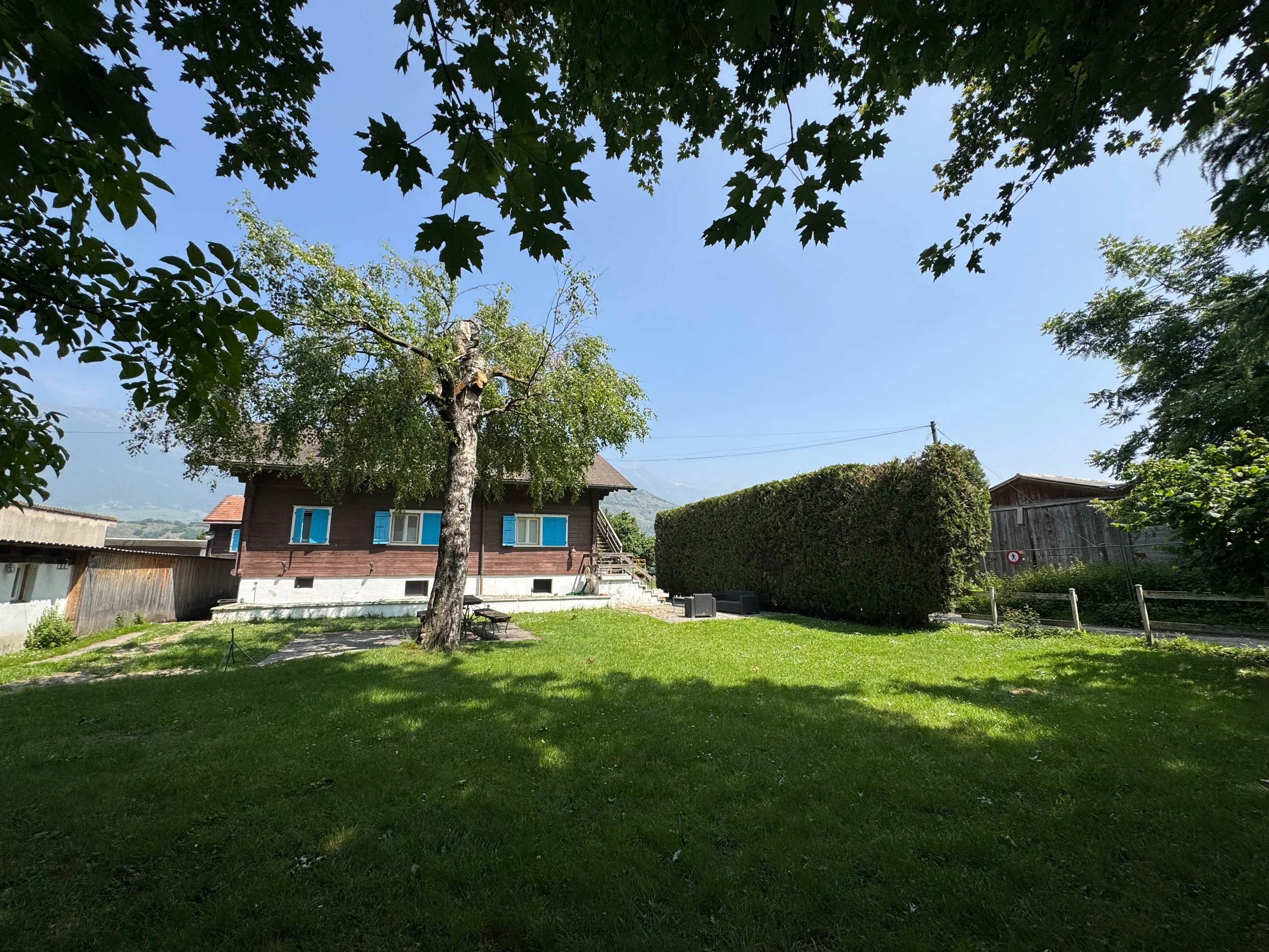 Maison en bois avec fenêtres bleues, entourée d'arbres et d'une pelouse verte, sous un ciel bleu clair. Gîte des Îles à Sion. Jardin, gazon, canapé de jardin.