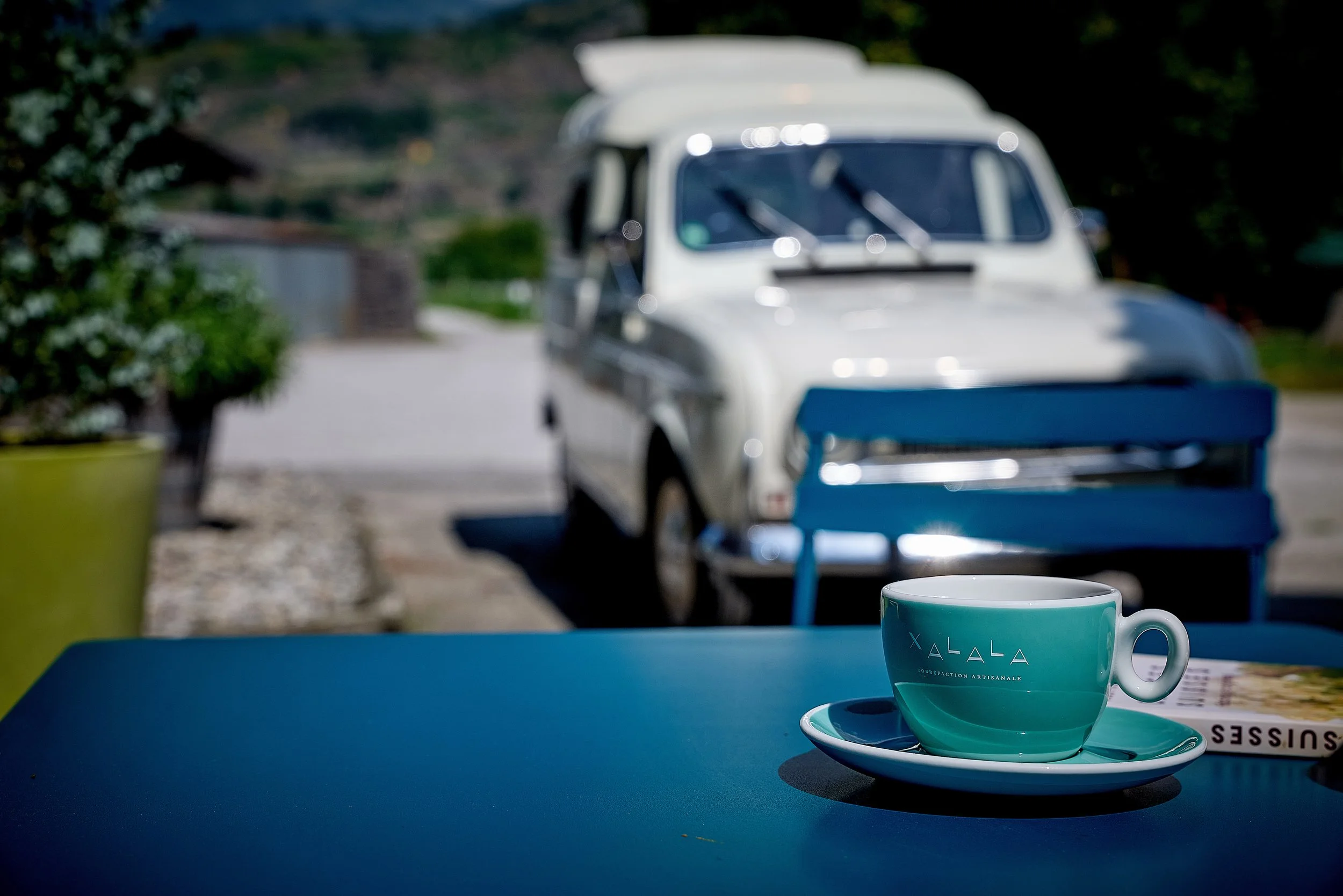 Une tasse turquoise avec une soucoupe sur une table bleu foncé, avec une petite voiture blanche vintage en arrière-plan et un livre à côté. Le tout est en extérieur, sous le soleil.