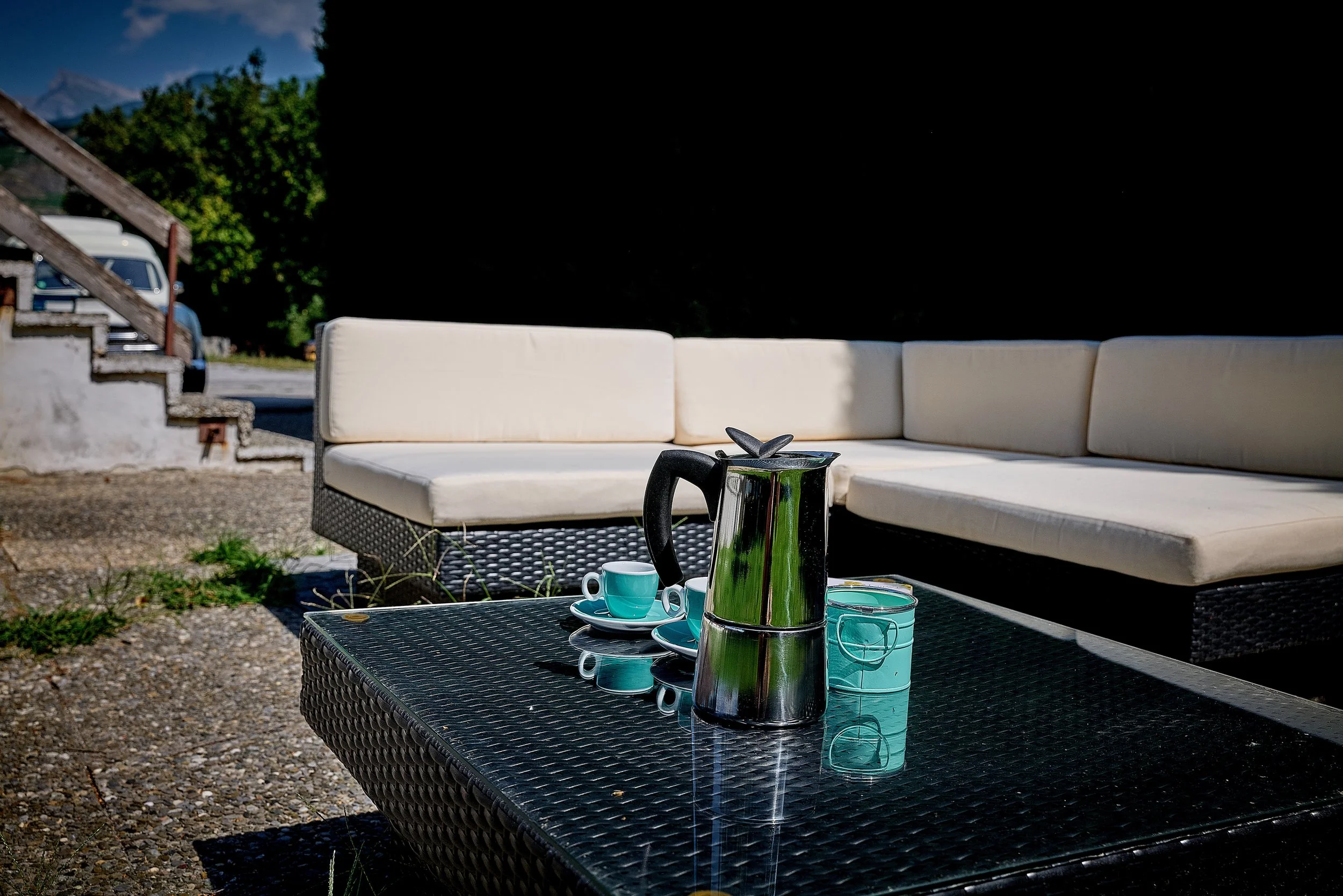 Table avec une cafetière, deux tasses à café, et une boîte en verre. En arrière-plan, un canapé blanc en extérieur et des voitures dans une zone en plein air.