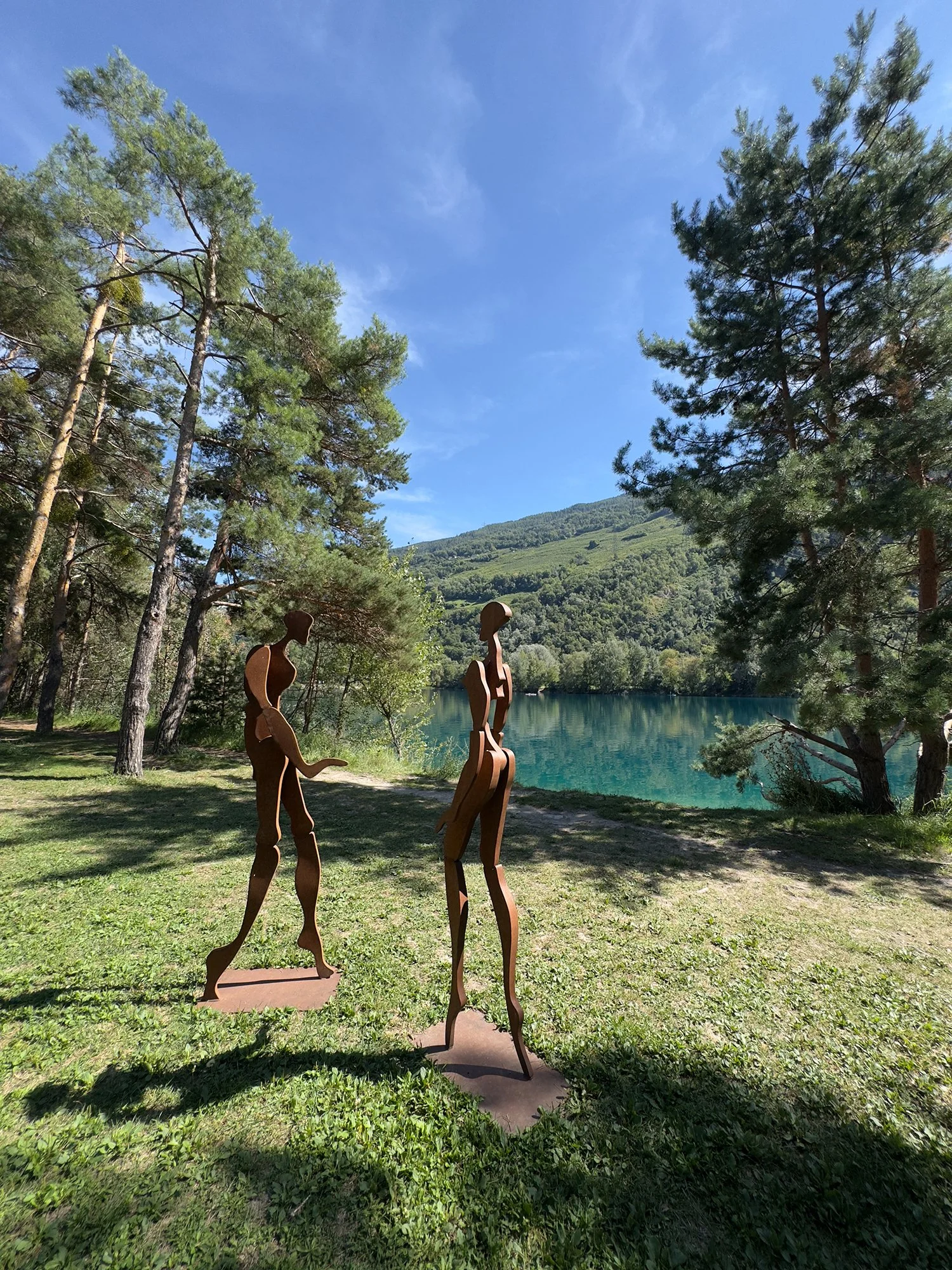 Deux sculptures en métal représentant deux figures humaines dans un paysage naturel avec des arbres, une étendue d'eau et une montagne en arrière-plan, sous un ciel bleu avec quelques nuages.