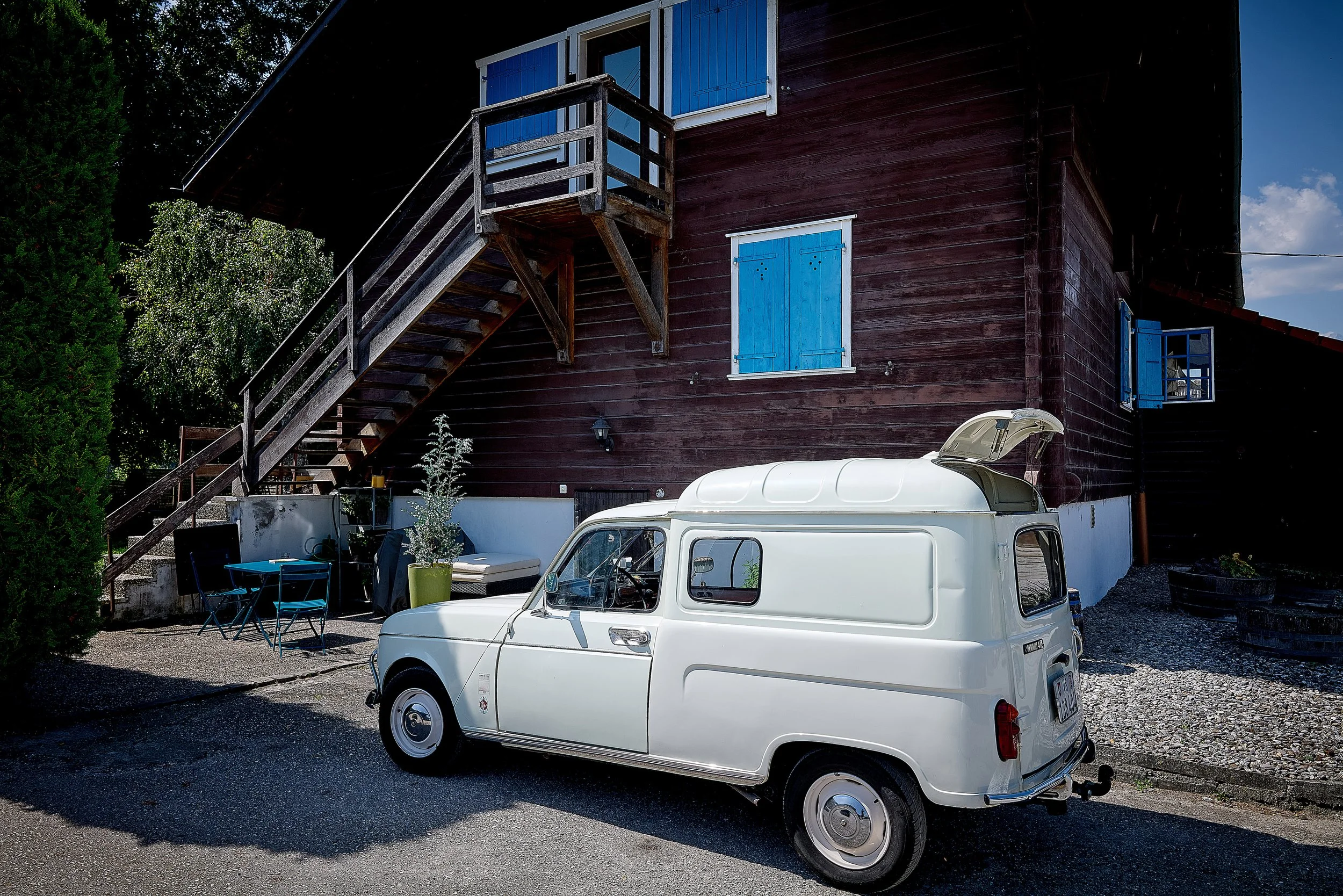 Une petite voiture blanche stationnée devant une maison en bois sombre avec des volets bleus. La maison a un escalier en bois menant à un balcon. Il y a une table avec des chaises et un fauteuil à côté du mur, ainsi que quelques plantes en pot.
