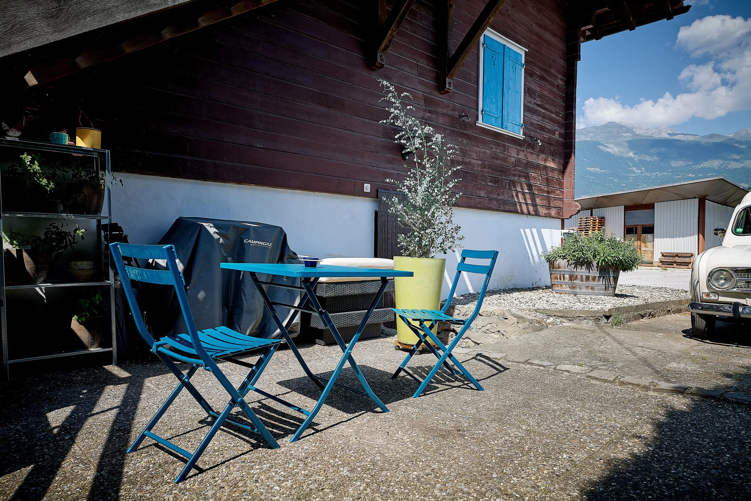 Terrasse extérieure avec deux chaises et table bleues, une plancha recouverte d'une housse, des plantes aromatiques, un bâtiment en bois, un véhicule ancien, et un paysage alpin en arrière-plan.