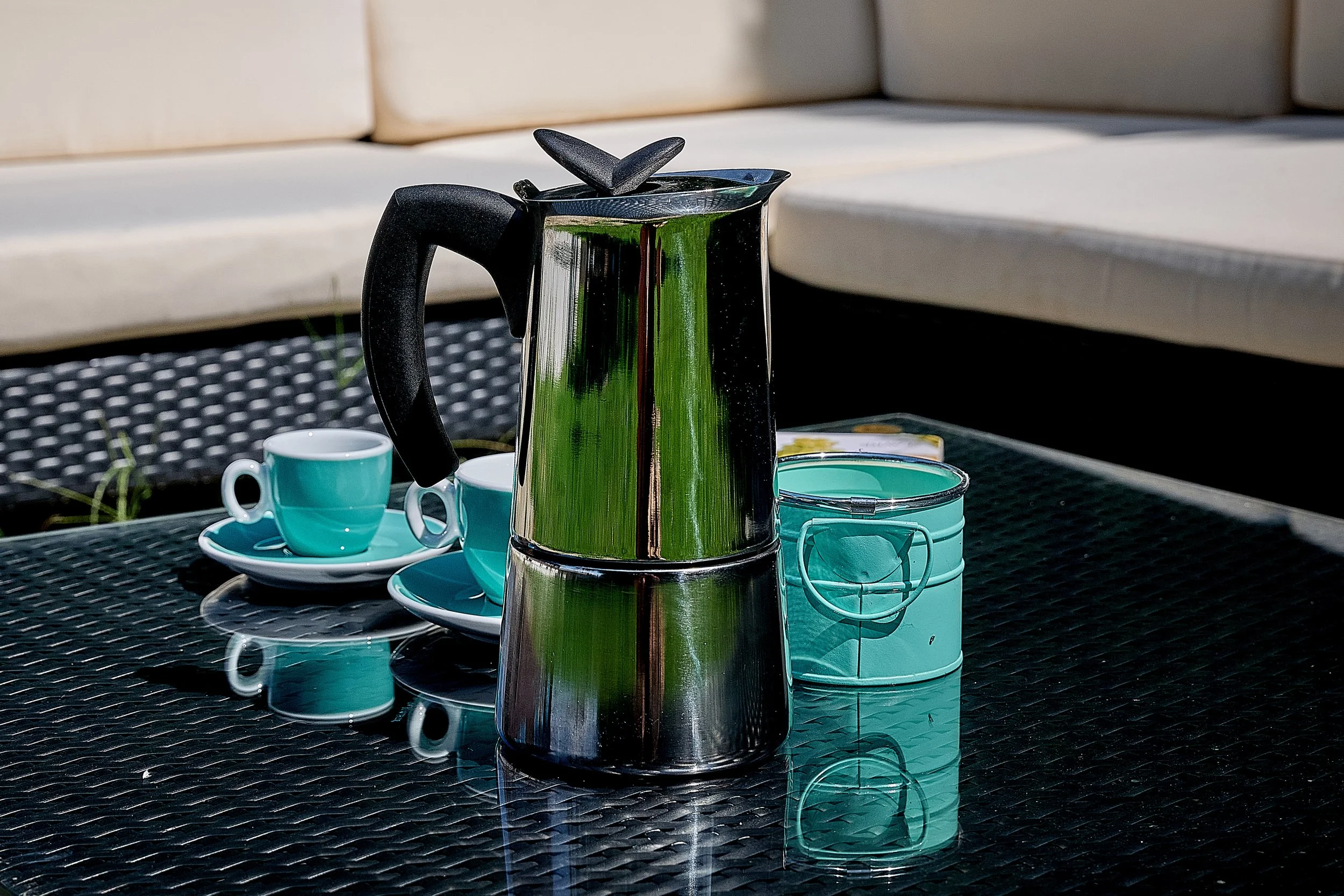 Une cafetière en acier inoxydable, deux tasses bleues avec soucoupes, et une boîte en métal sur une table en verre noir à l'extérieur. Xalala Chamoson café.