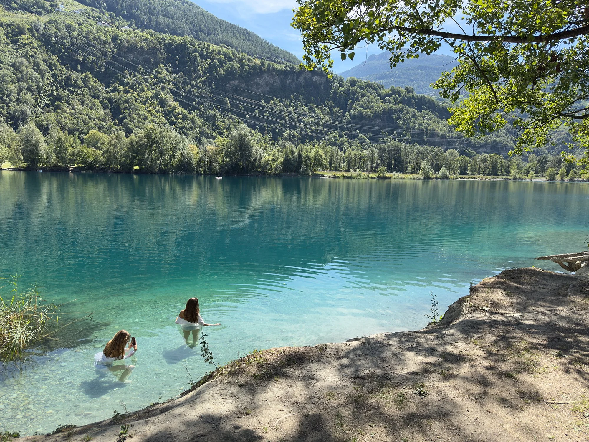 Deux femmes nagent dans un lac turquoise entouré de montagnes verdoyantes, avec des arbres ombrageant partiellement la scène. La surface de l'eau est calme et claire, reflétant le paysage environnant.