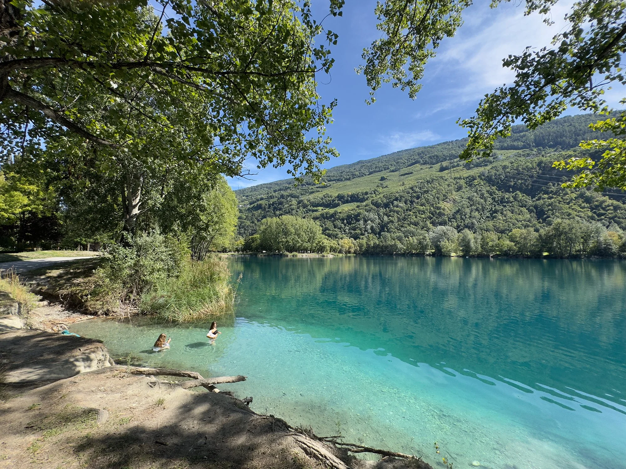 Un lac entouré d'arbres verts et de montagnes sous un ciel bleu, avec deux personnes nageant dans l'eau. Au Domaine des Îles de Sion, en Valais (Suisse). Une plage de galets au bord d'un lac turquoise entouré de végétation et de montagnes, avec un ci