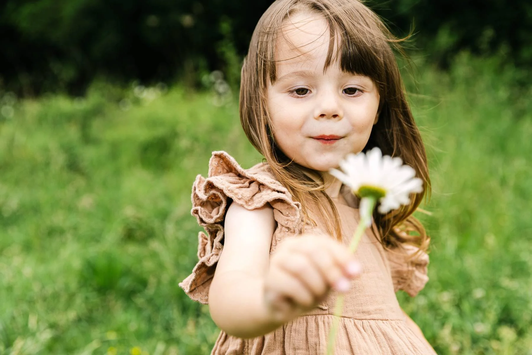 A young girl with brown hair and a beige dress holding a white daisy flower outdoors on a grassy field.