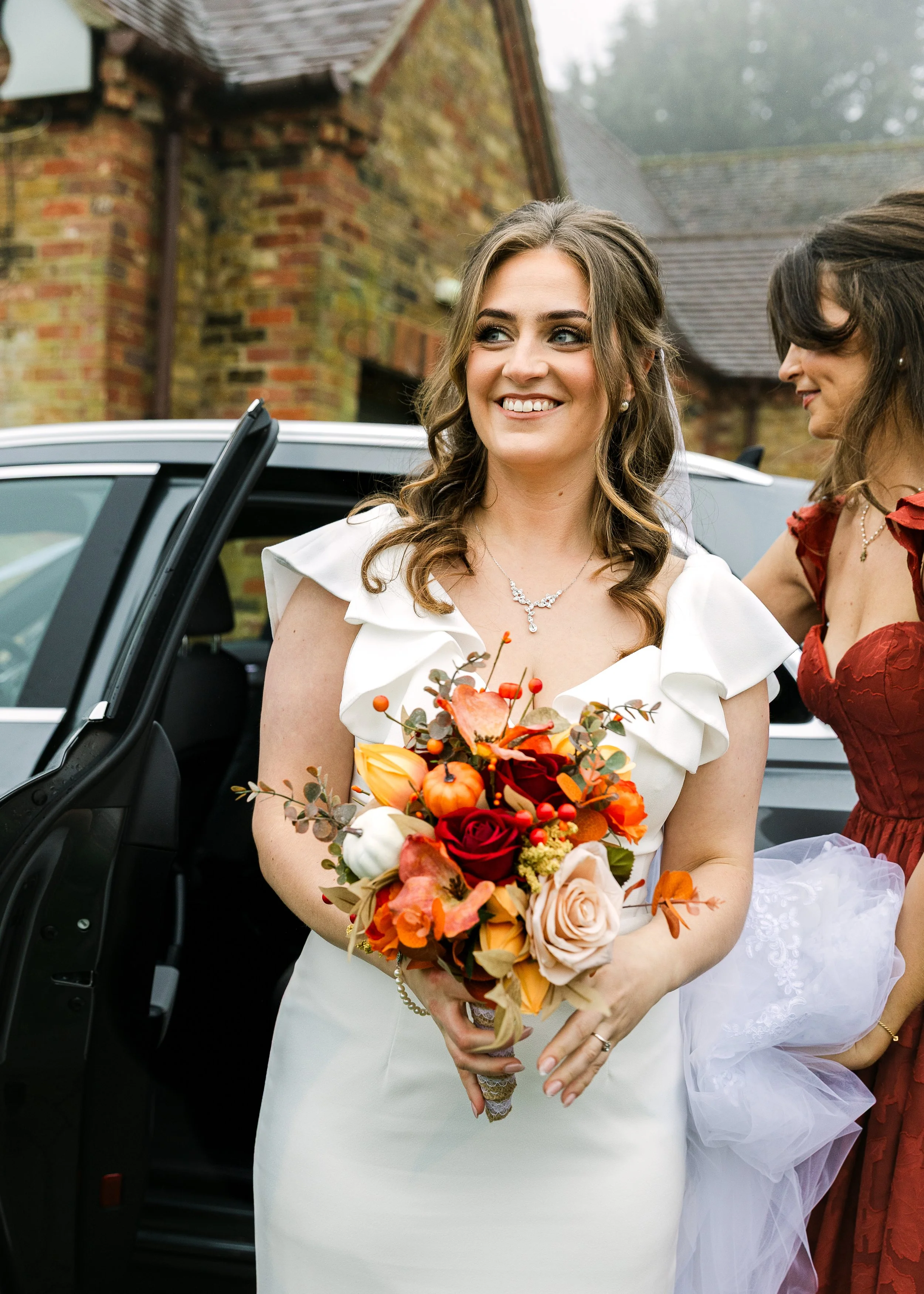 Autumn wedding in Bedfordshire, the bride is stepping out of the car with a autumnal themed boquet in her hands. She is wearing an ivory fitted wedding dress with frills around the capped sleeves and neckline