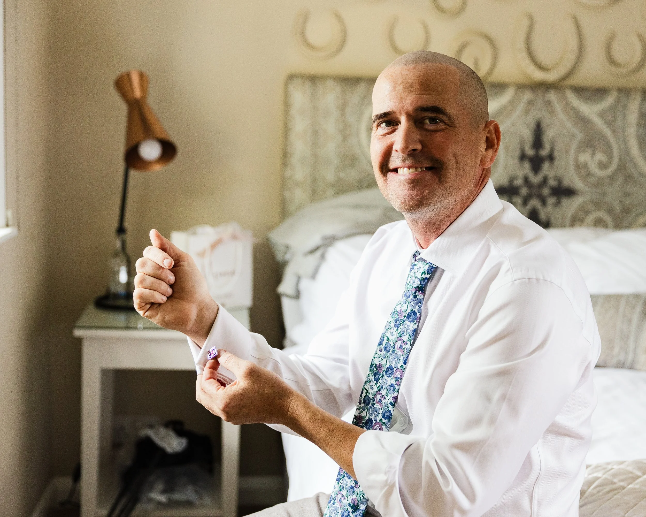 A man with a shaved head and light stubble, smiling, is sitting on a bed in a hotel room, putting on a cufflink on his white dress shirt sleeve, wearing a patterned blue tie. The background shows a bedside table, a lamp, and decorated wallpaper.