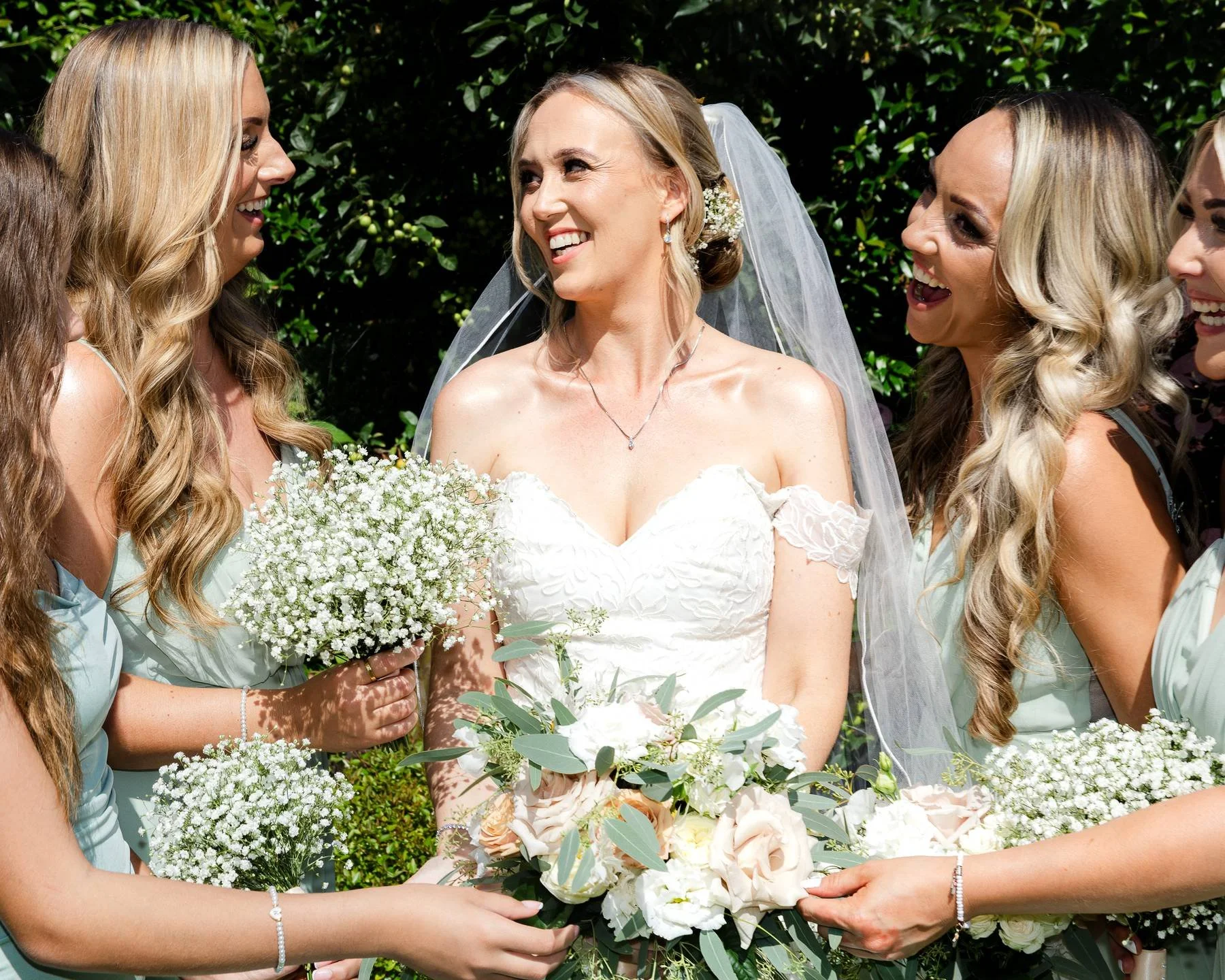 A bride in a white wedding dress surrounded by bridesmaids holding bouquets of white flowers, all smiling and looking joyful outdoors.