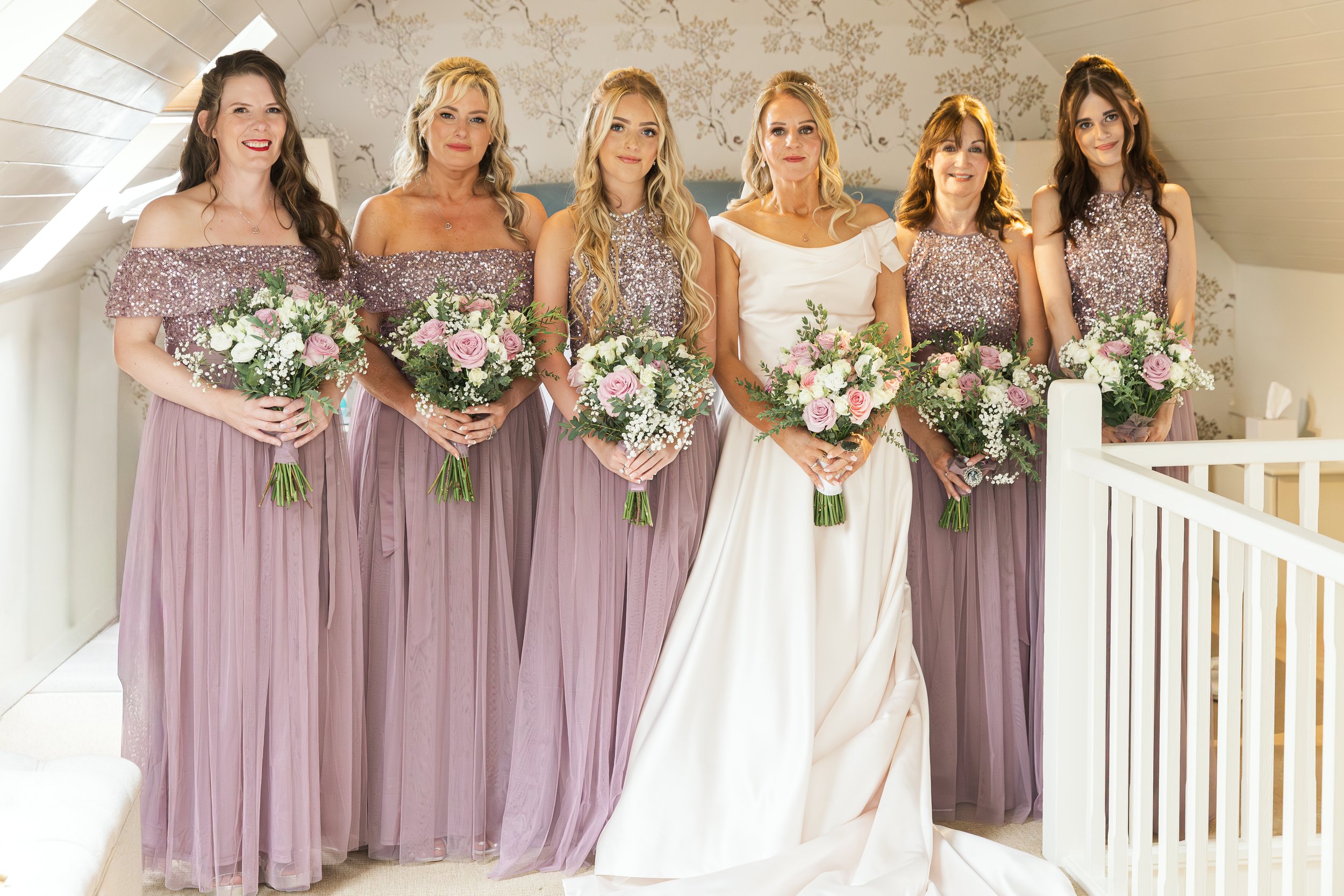A bride and six bridesmaids standing together at a wedding, holding bouquets of pink and white flowers, inside a decorated room with a sloped ceiling.