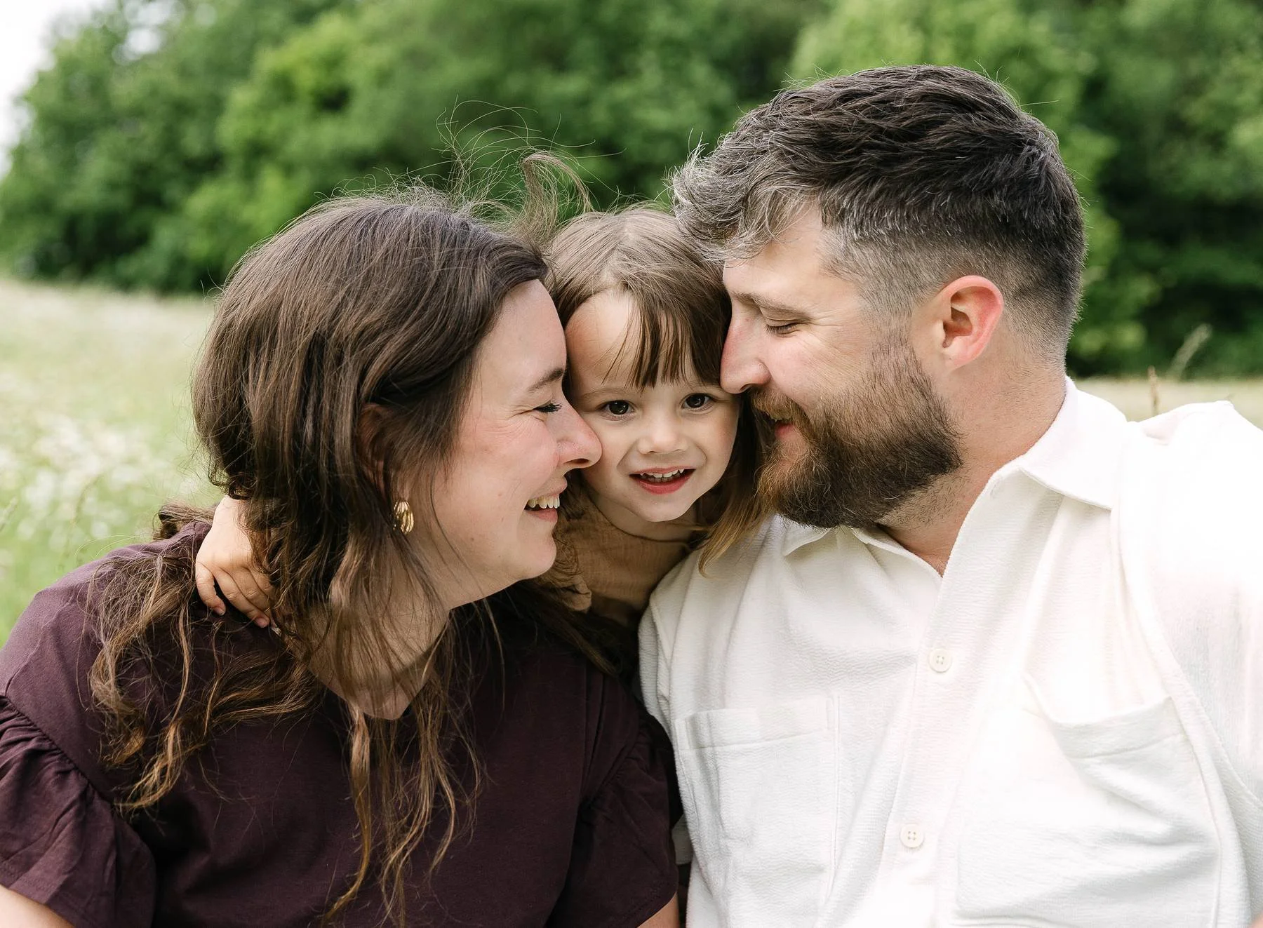 A family of three, smiling and touching foreheads, outdoors in a green field.