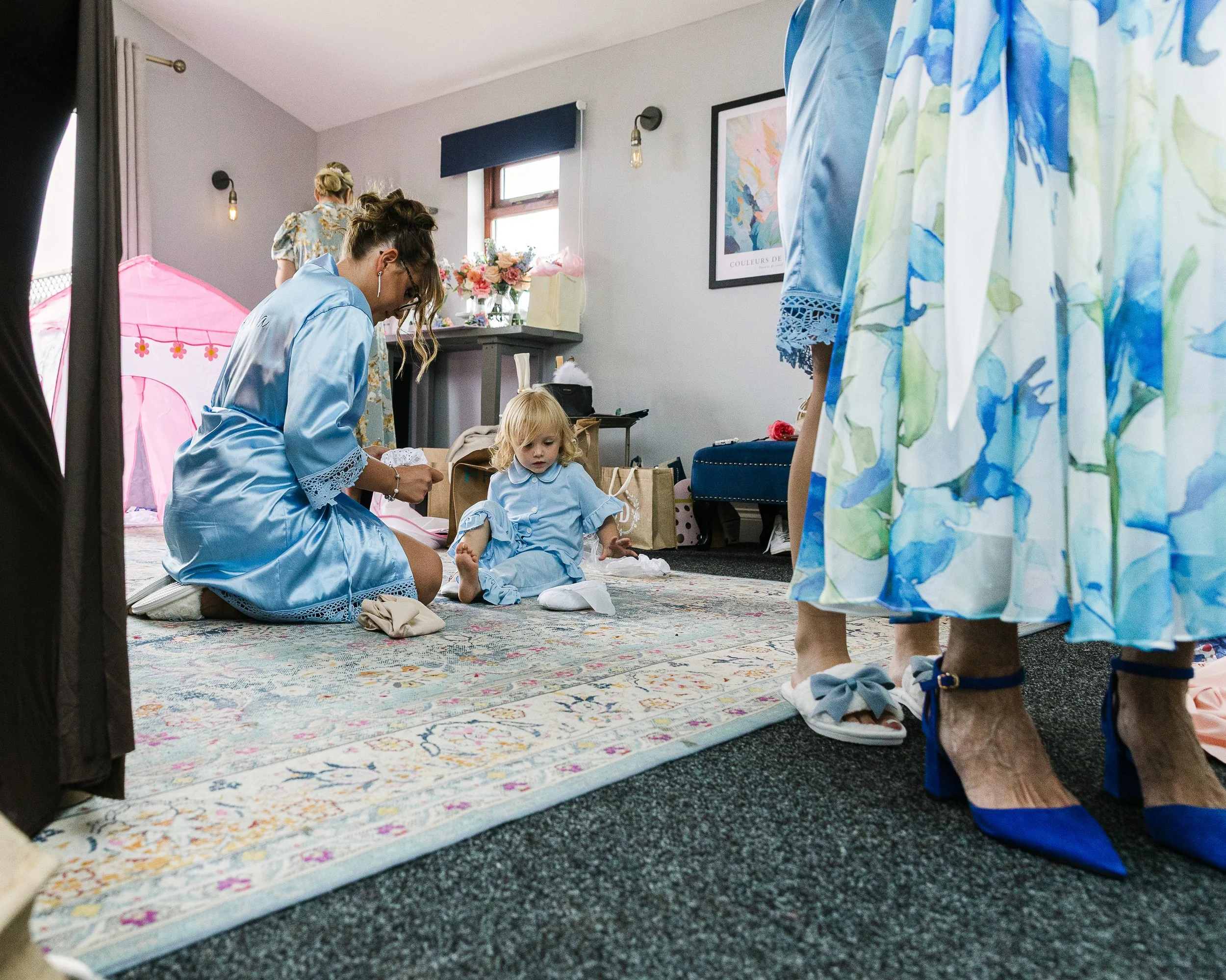 Young flower girl sitting on the floor having her socks put on by an older bridesmaid with two sets of feet in the foreground