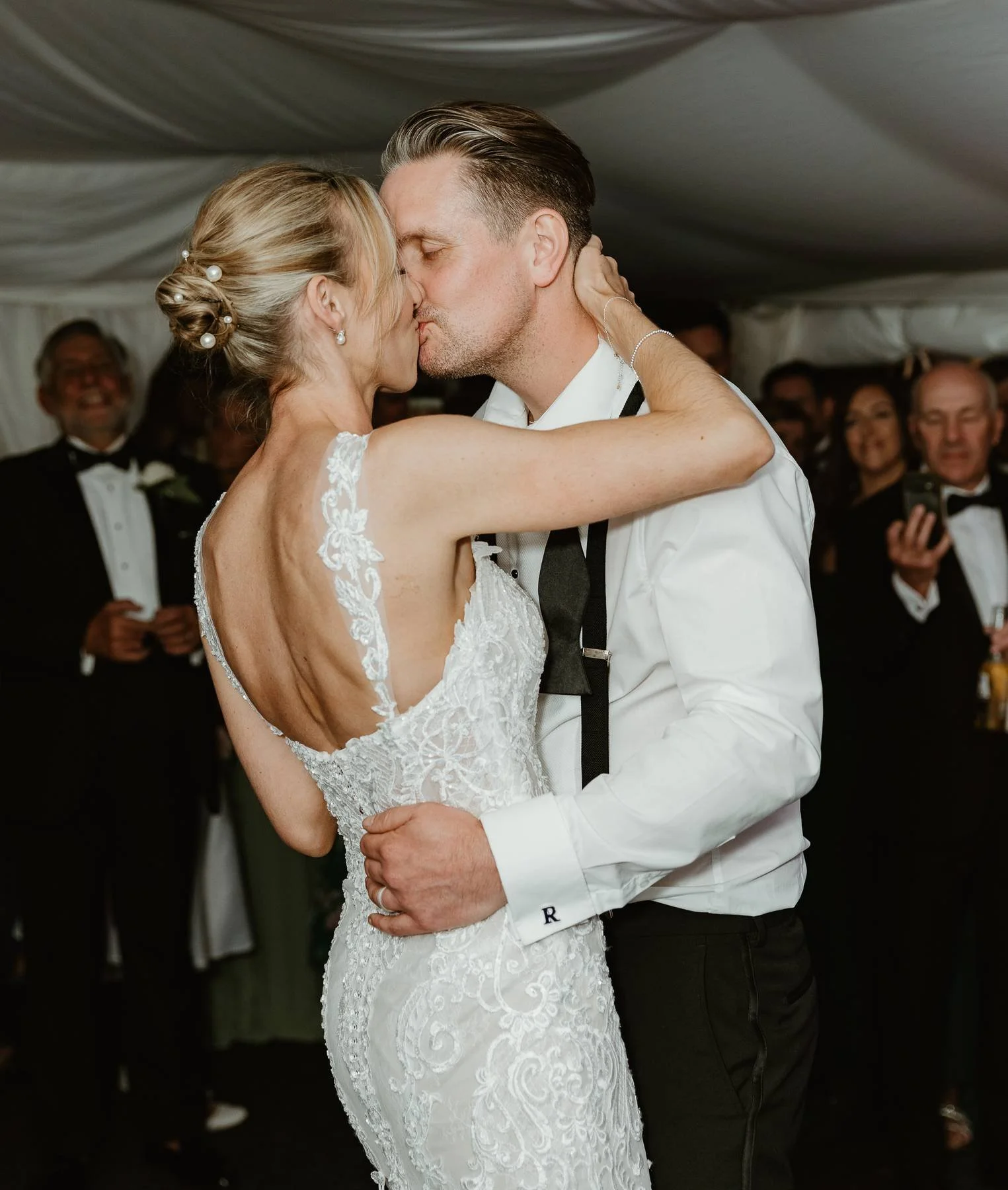 A bride and groom kiss during their wedding dance inside a decorated tent, surrounded by guests in formal attire.