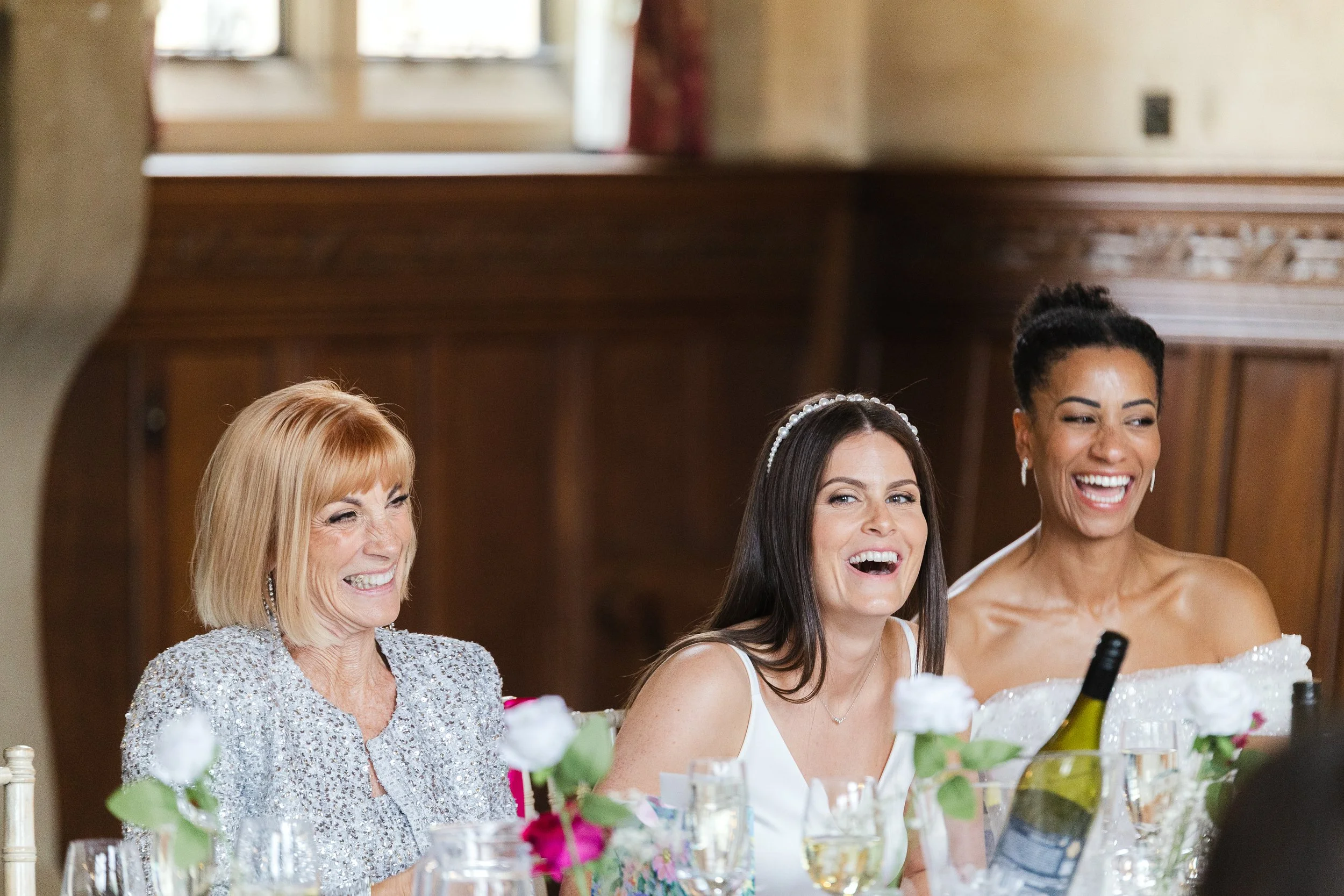 Three women at a formal event, laughing and smiling, with a wooden-paneled background, floral arrangements, and wine bottles on the table.