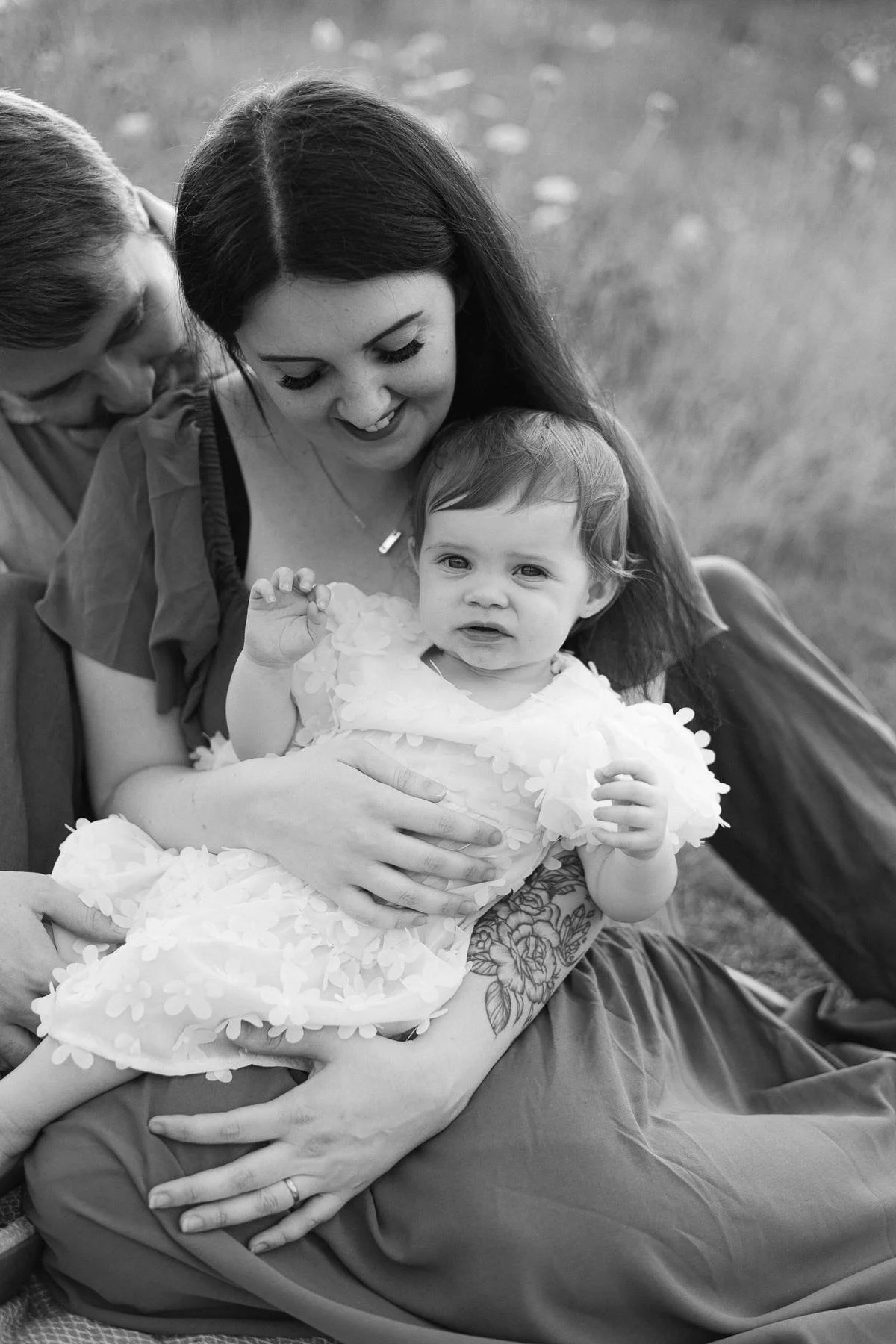 A woman holds a young girl in her arms, sitting outdoors on grass, both smiling and looking at the camera. Another person leans in from the side, partially visible. The girl wears a dress with floral appliqué and has short hair. The woman has long da