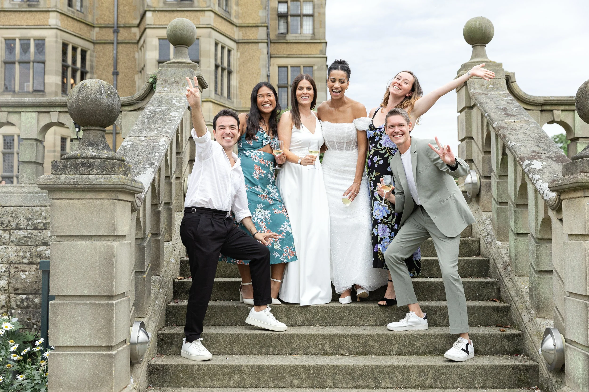 Group of friends celebrating at a wedding outside on stairs, holding drinks, smiling and posing happily.