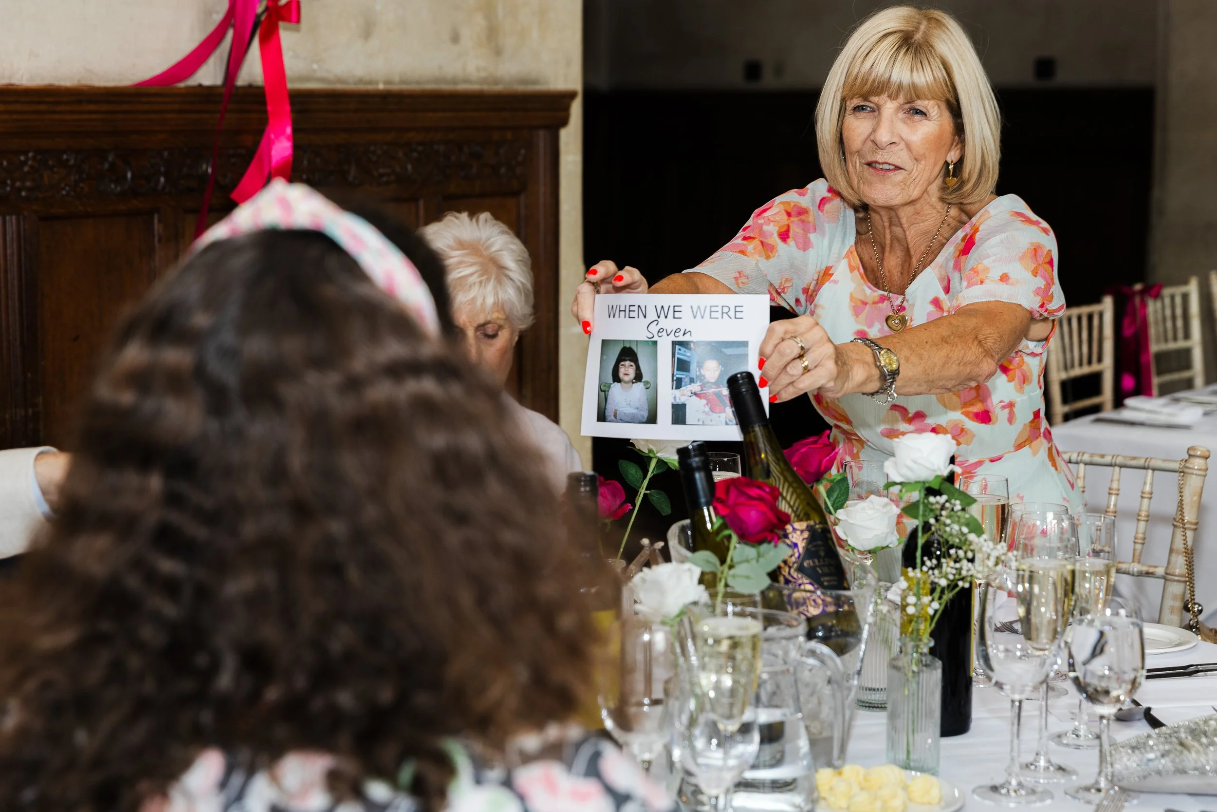An elderly woman at a formal event holding a sign that says "When We Were Seven" with two childhood photos. She is standing at a decorated table with flowers, wine bottles, and glasses, and speaking to a group of seated people.