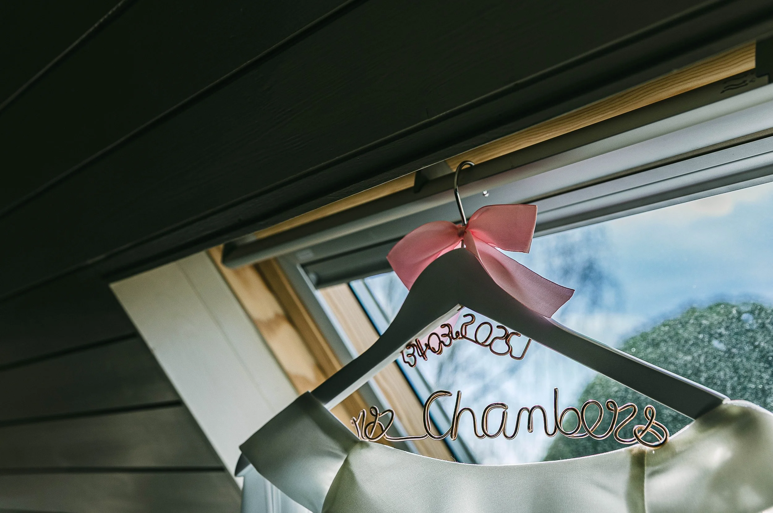 Close-up of a white dress on a hanger with a pink bow, hanging near a window with a green outdoor view.
