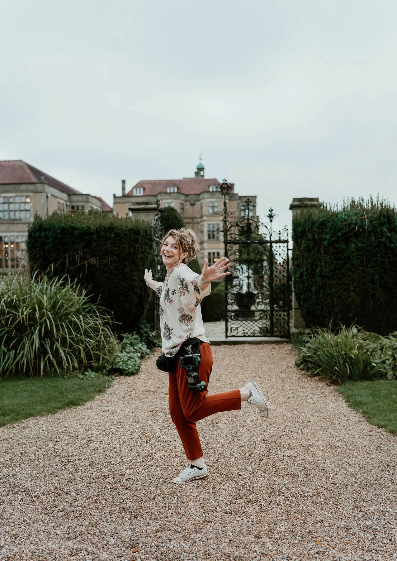 A woman with a camera hanging from her waist, jumping joyfully on a gravel path with greenery and a wrought iron gate behind her, in front of an elegant building.