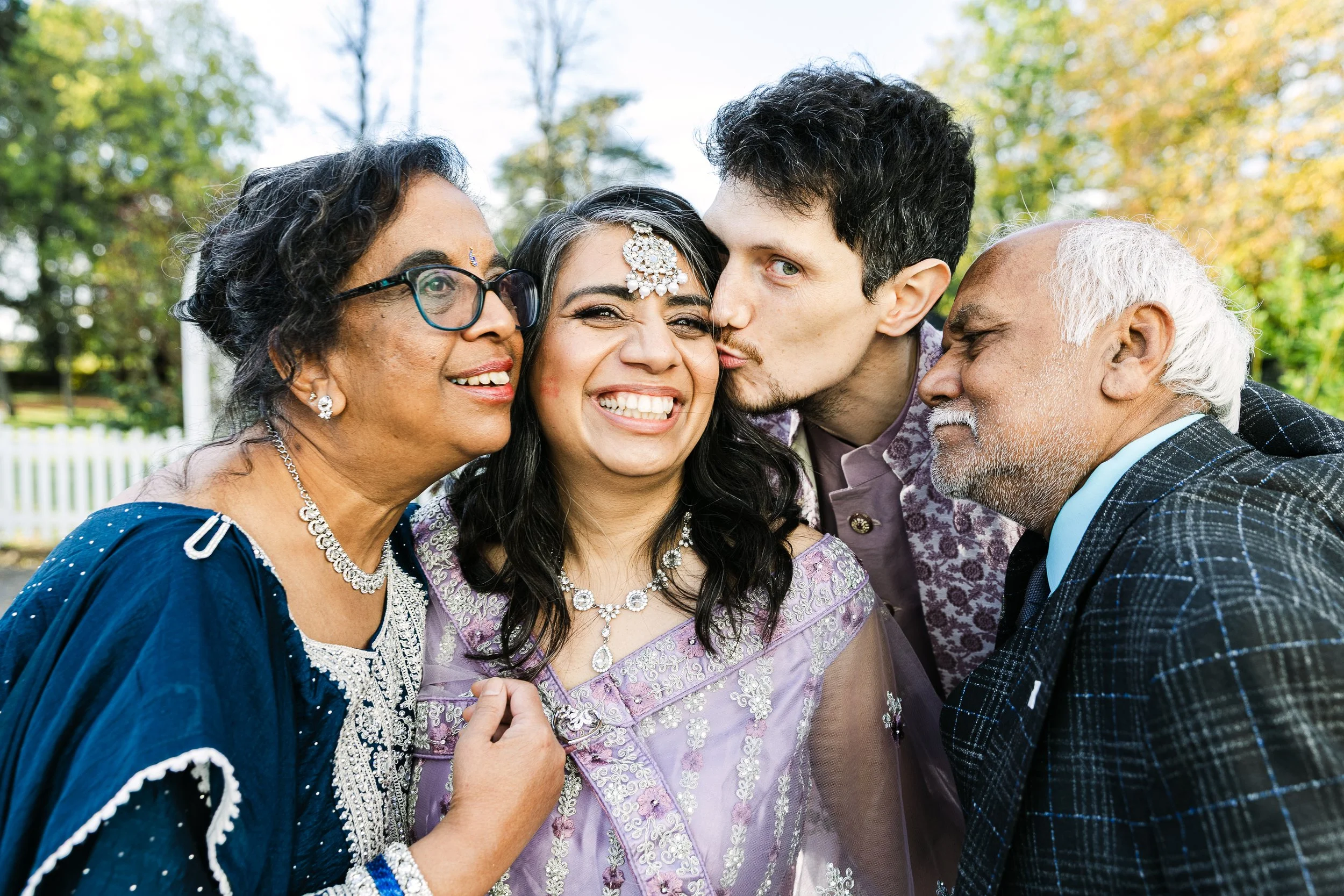 Asian bride with her parents and husband, all leaning in close smiling at the bride. Groom is kissing her cheek