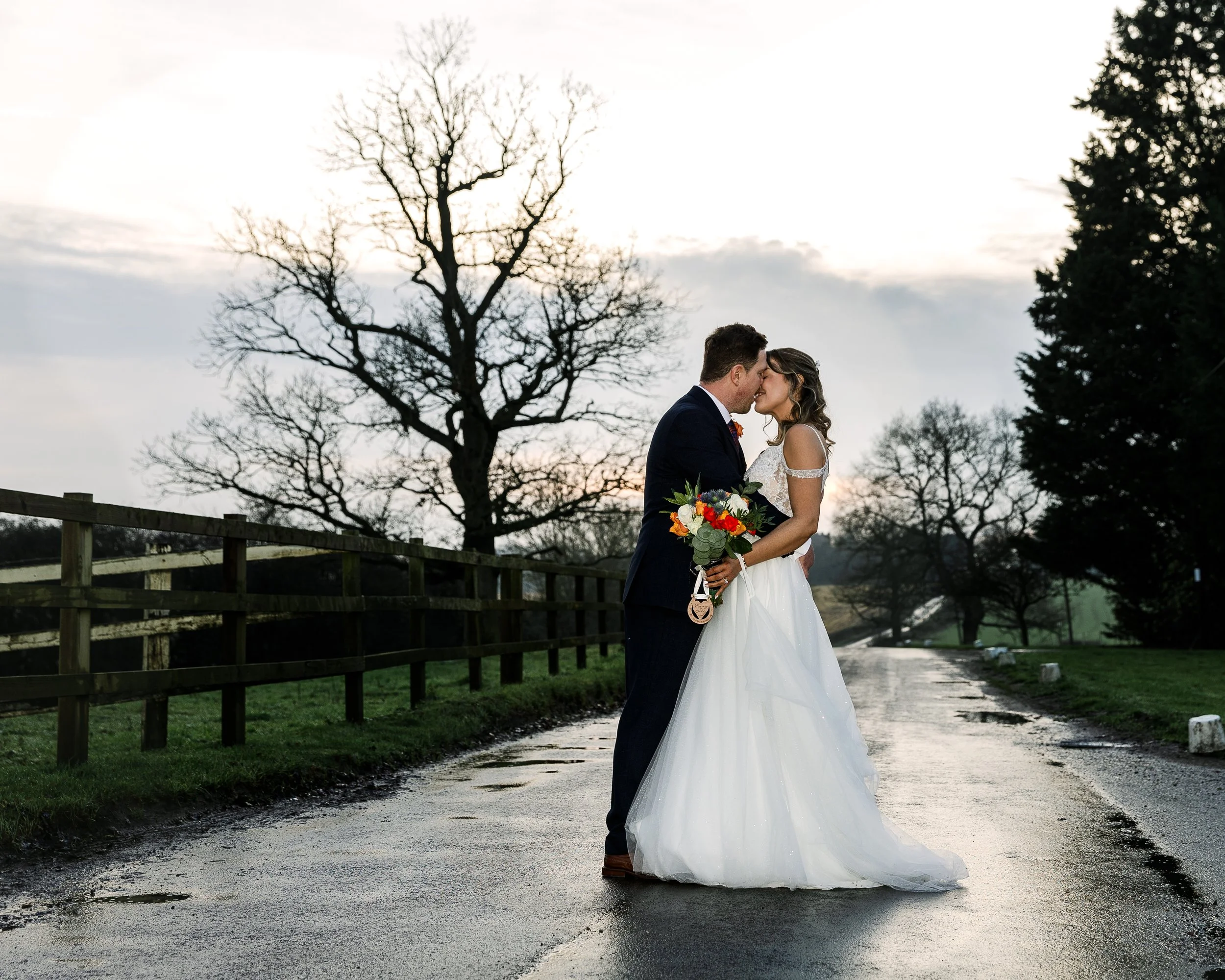 A bride and groom sharing a kiss on a wet road during sunset, with leafless trees in the background, the bride holding a bouquet of colorful flowers.