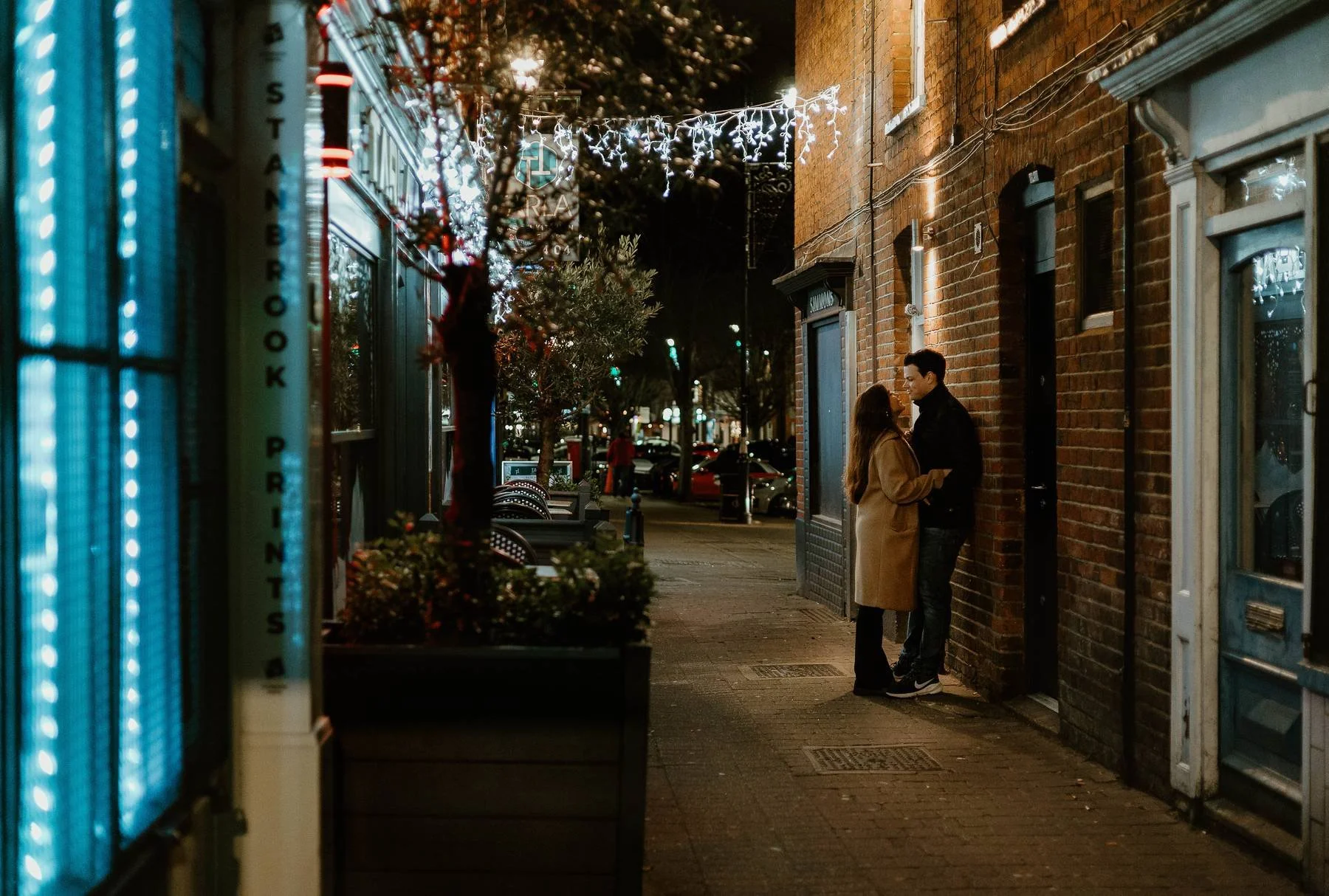 A couple stands close together on a sidewalk at night, leaning against a brick building, with lights hanging overhead in a festive setting.