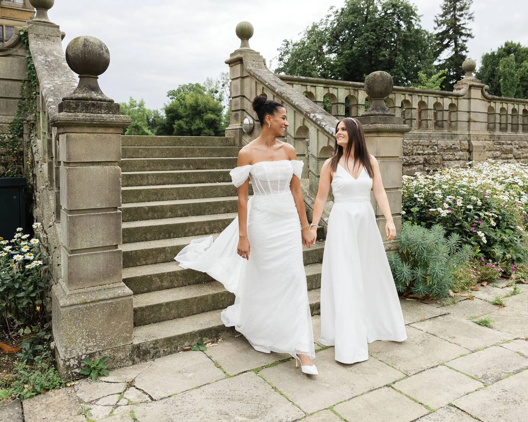 Two women in wedding dresses holding hands and smiling at each other outdoors near stone steps and flowers.