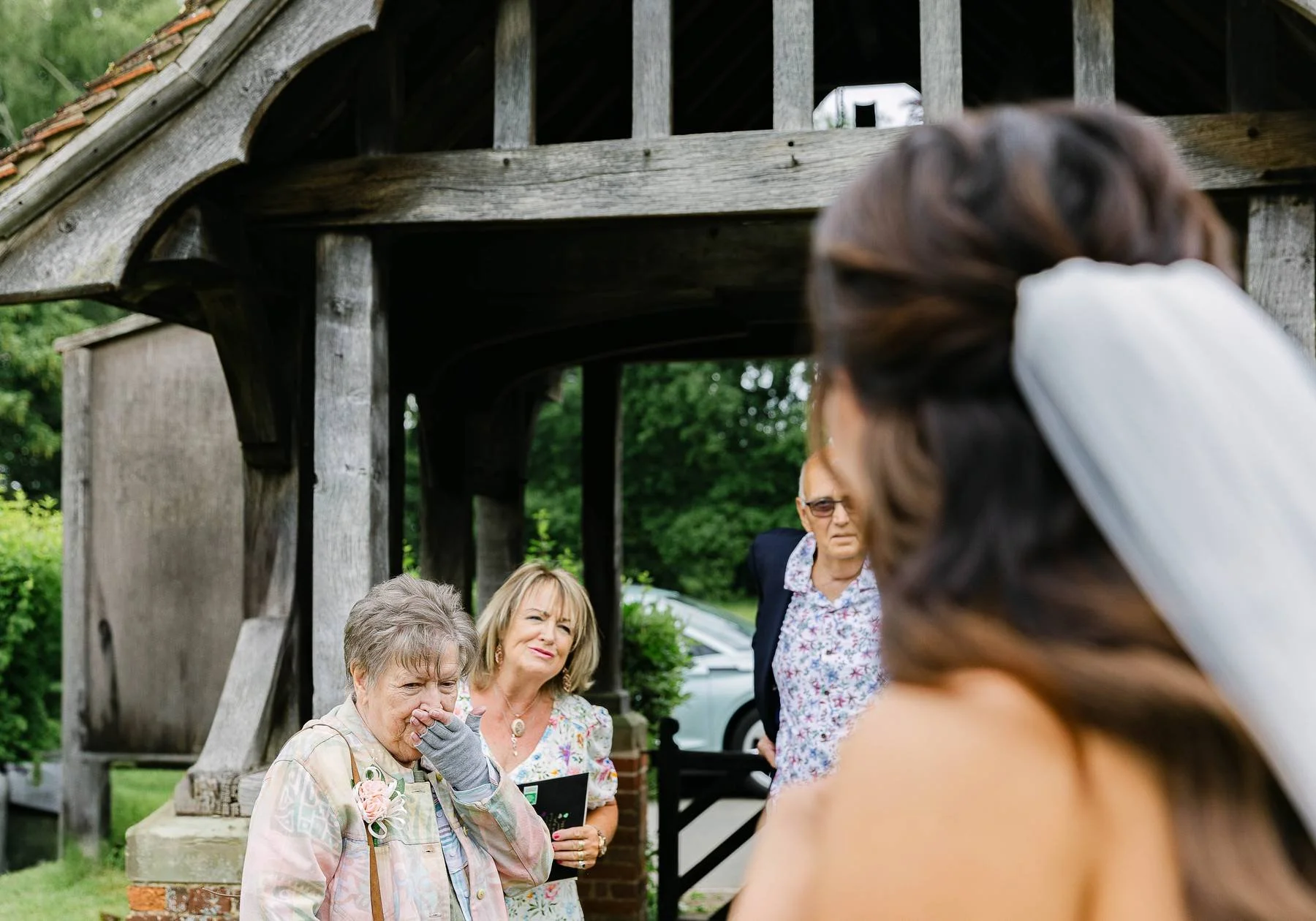 A bride in a wedding dress with a veil is facing a group of three older women outdoors. One woman is crying and holding her nose, another woman is smiling and holding a photo, and a man in glasses and a floral shirt is standing behind them under a wo