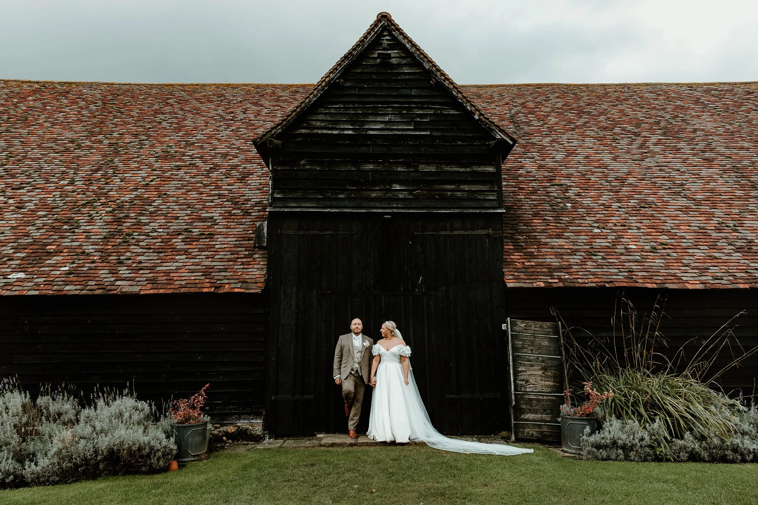Bride and groom holding hands standing in front of a large black barn with a weathered, shingled roof on a cloudy day.