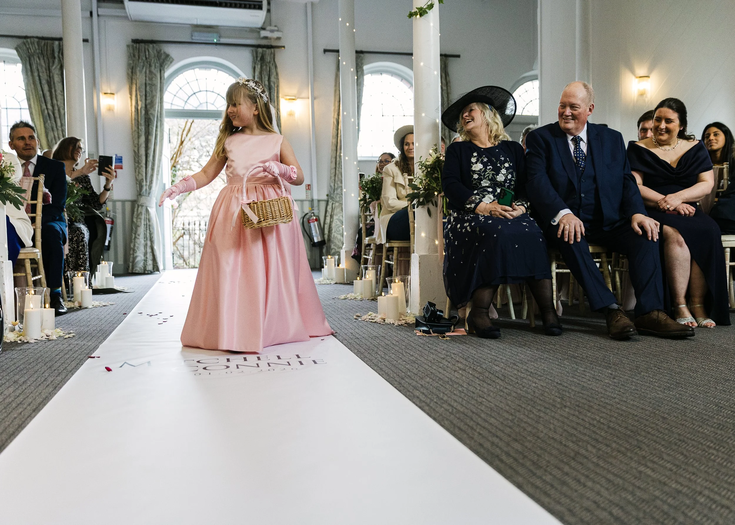 Young girl in a pink dress, wearing pink gloves and holding a small basket, walking down an aisle at a wedding ceremony. Guests are seated on either side, some smiling and taking photos, with candles and decorations along the aisle.
