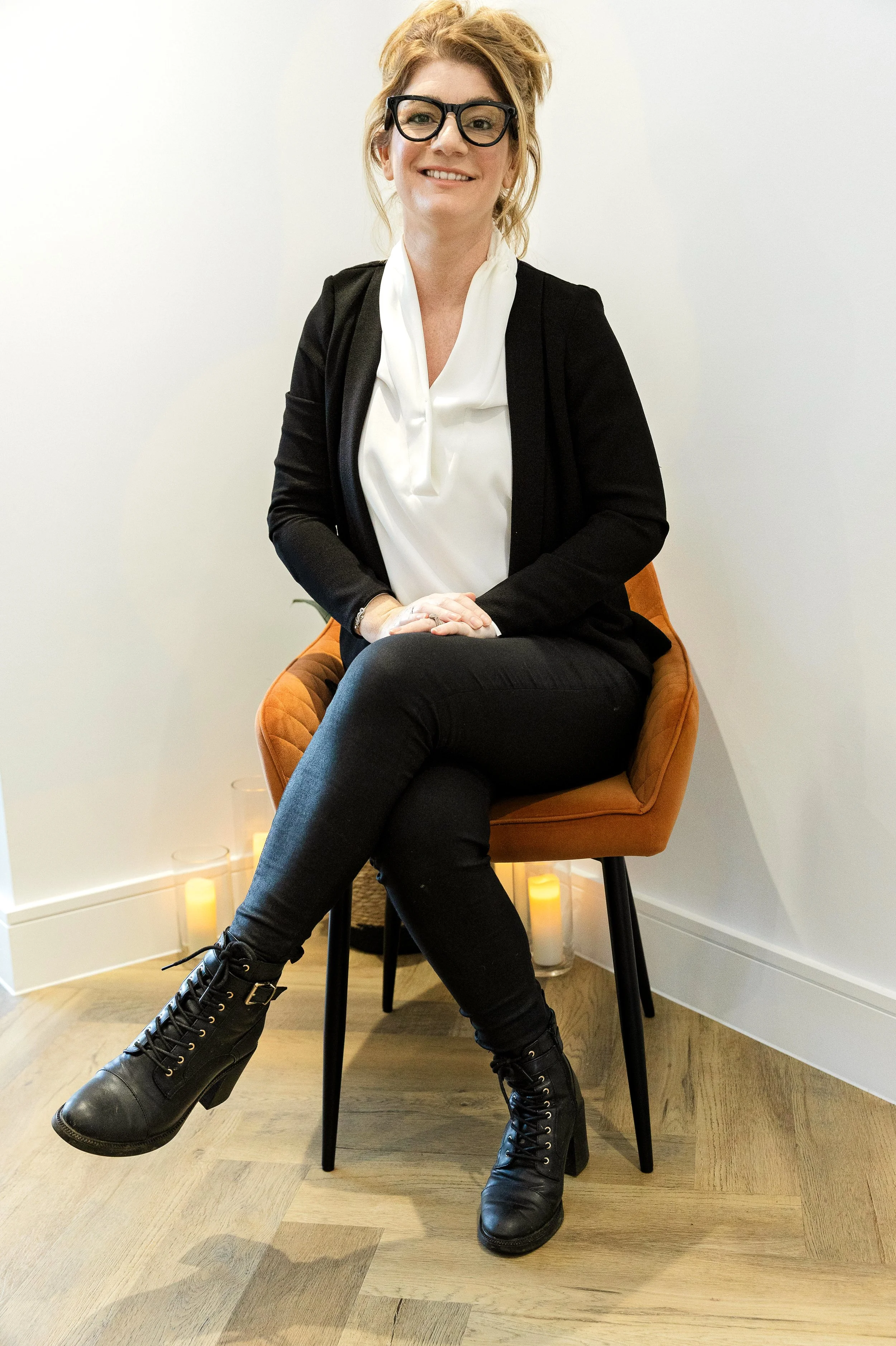 A woman with light skin, curly blonde hair, and glasses, smiling while sitting on a brown chair in a minimalistic room with candles on the floor.
