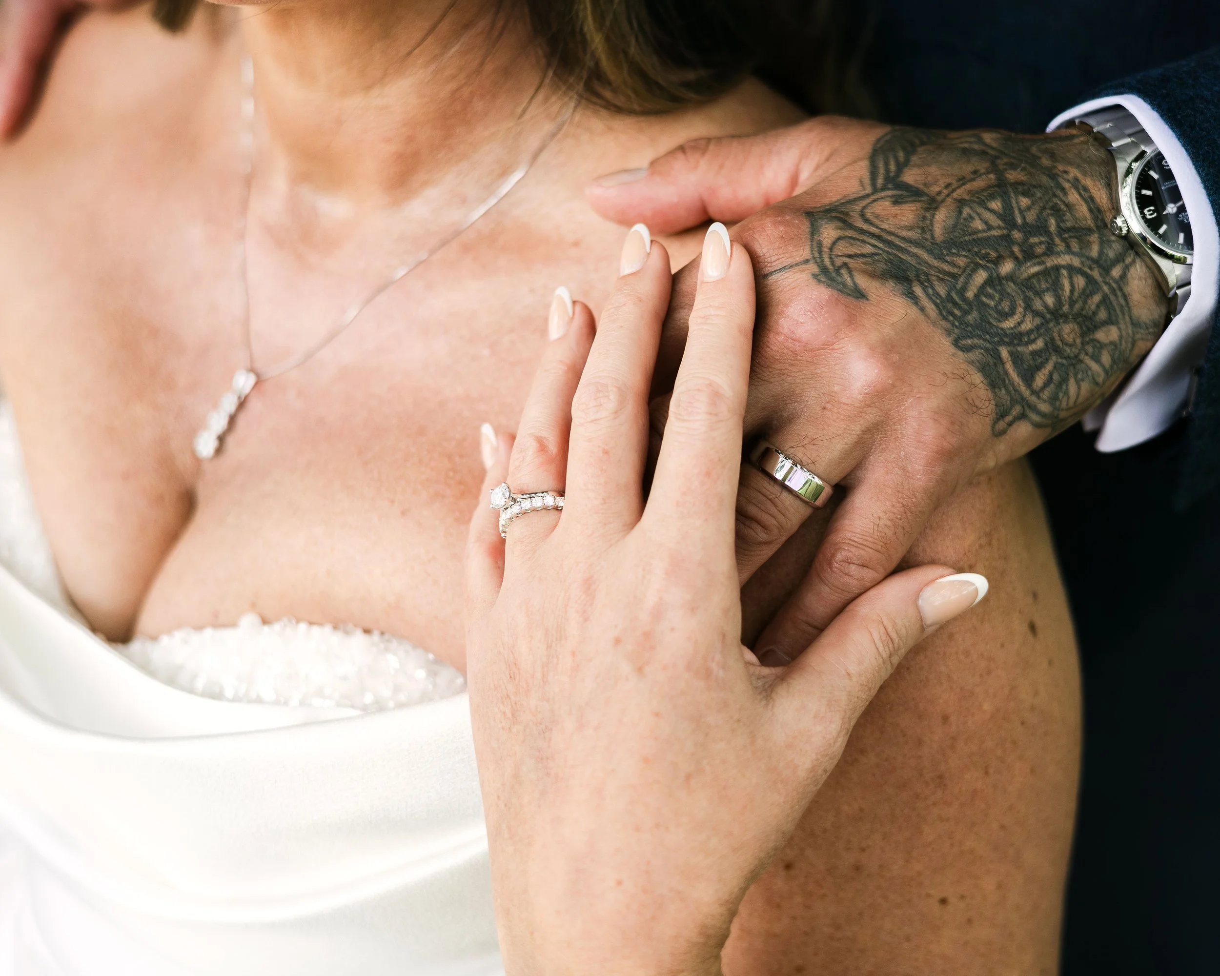 Close-up of a couple's hands showing wedding rings, with one hand on the woman's shoulder and the other on her back. The woman is wearing a white strapless dress and the man has a tattooed arm with a wristwatch.