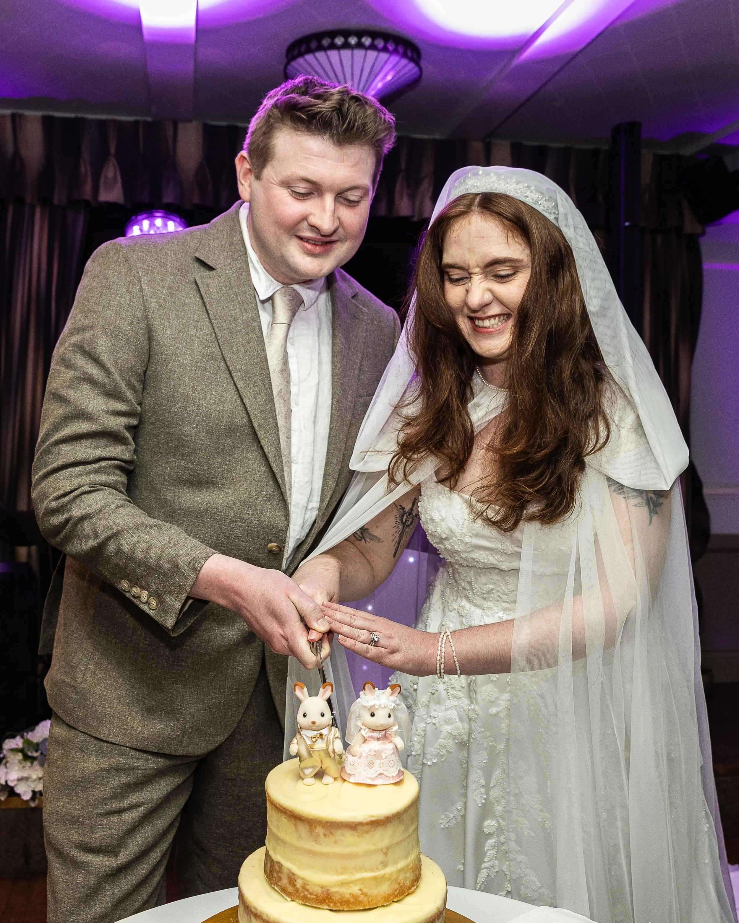 A bride and groom cut a wedding cake with rabbit figurines on top, smiling and enjoying their special moment.