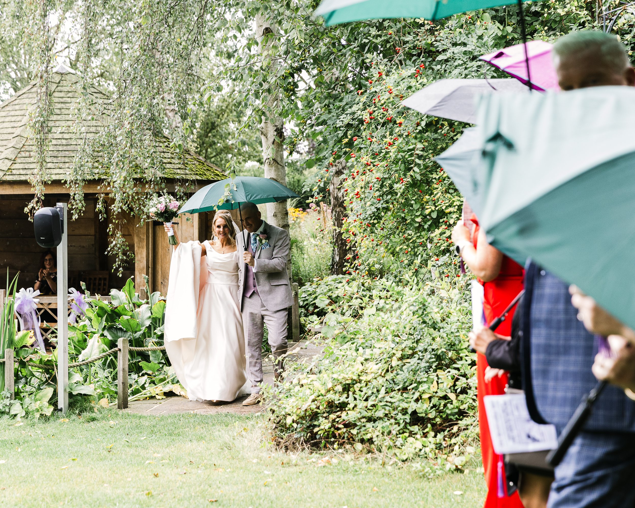 Bride and Groom at South Farm Hertfordshire walking under an umbrella toward a line of their wedding guests under umbrellas