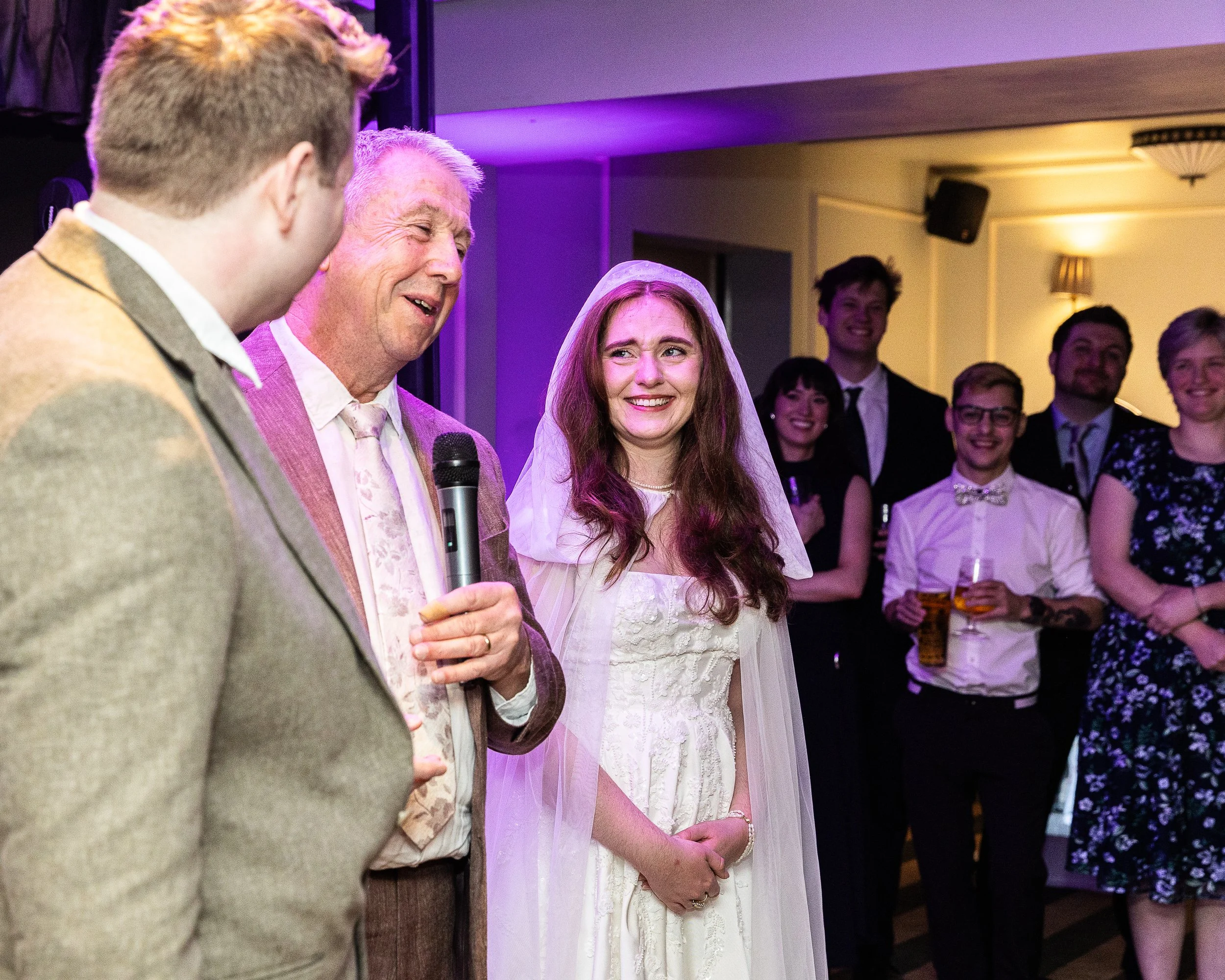 Father of the groom making a speech as the bride looks on with guests in the background on this wedding day in hertfordshire