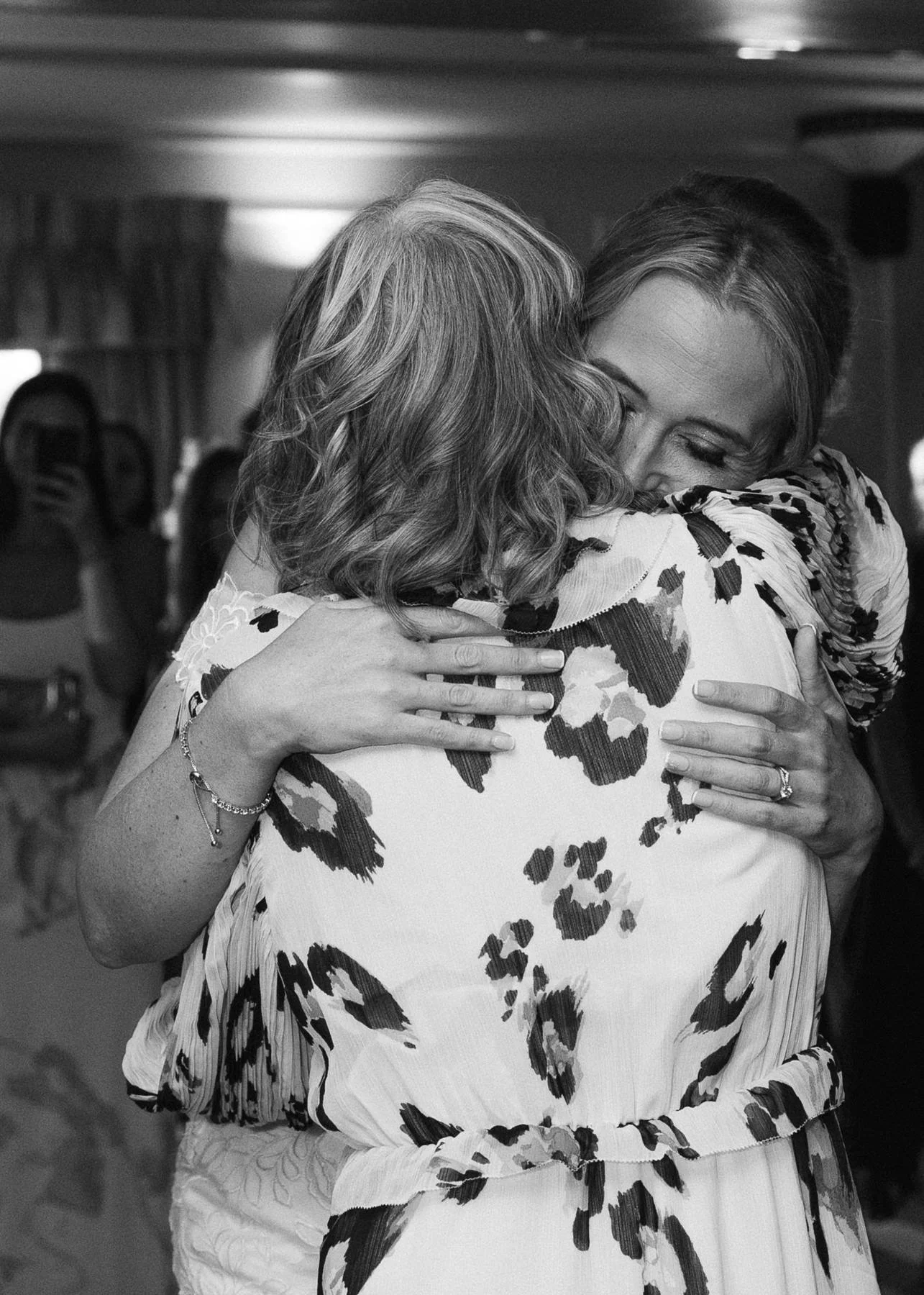 Two women hugging warmly, one with curly hair and the other with straight hair, in a moment of affection at an indoor gathering.
